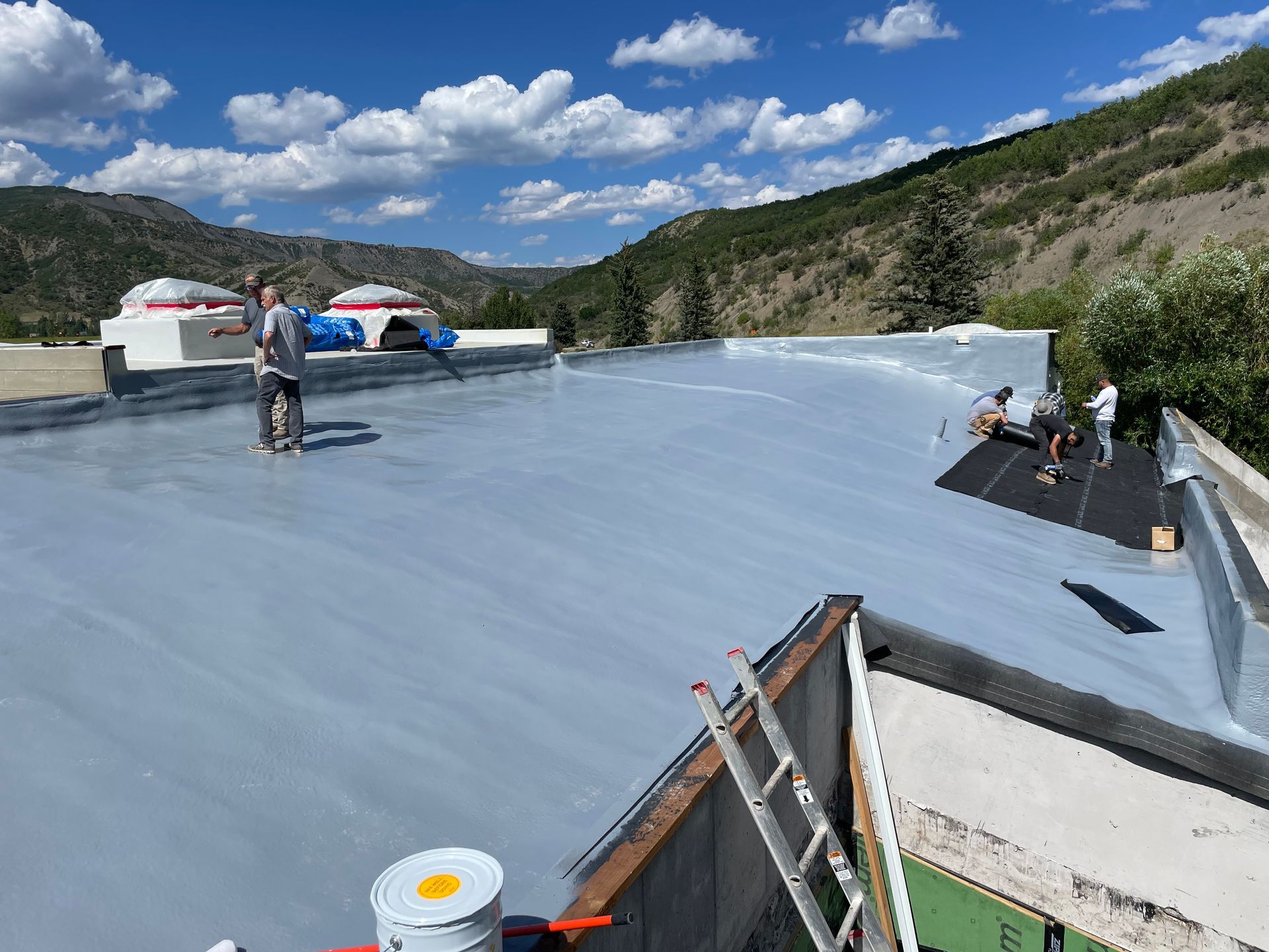 Workers applying coating to a flat roof, mountains in the background. Blue sky, grey coating, sunny day.