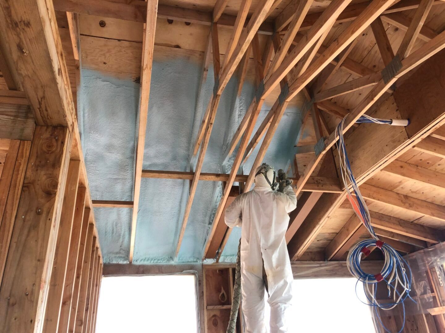 Person in protective suit spraying blue insulation onto a wooden ceiling during construction.
