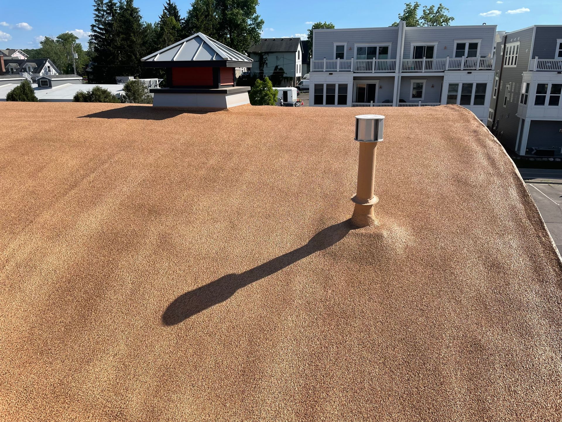 A flat roof covered in light brown mulch with a vent pipe and chimney in the sun.