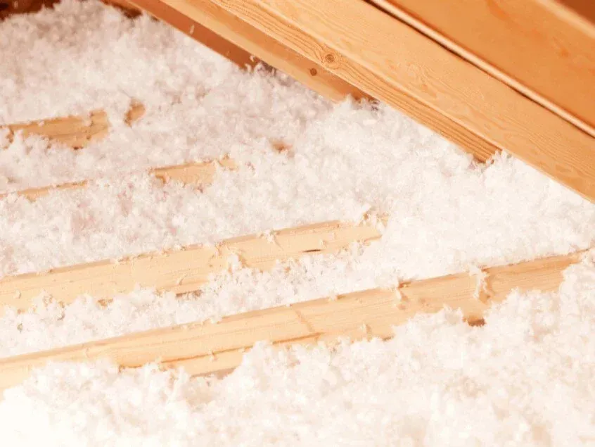 Attic with loose-fill insulation between wooden rafters. White insulation is spread.