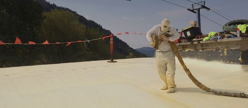 A worker in protective gear applies a thick foam coating to a large surface outdoors.