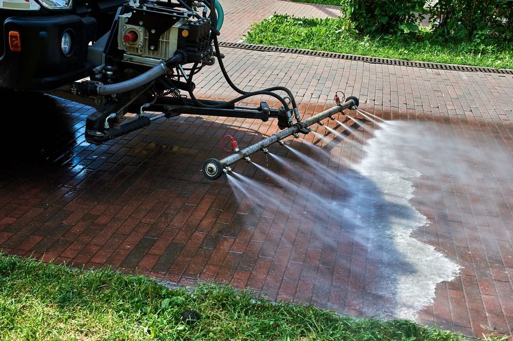 A vehicle-mounted spray bar cleans a brick patio with pressurized water, removing dirt from the surface.