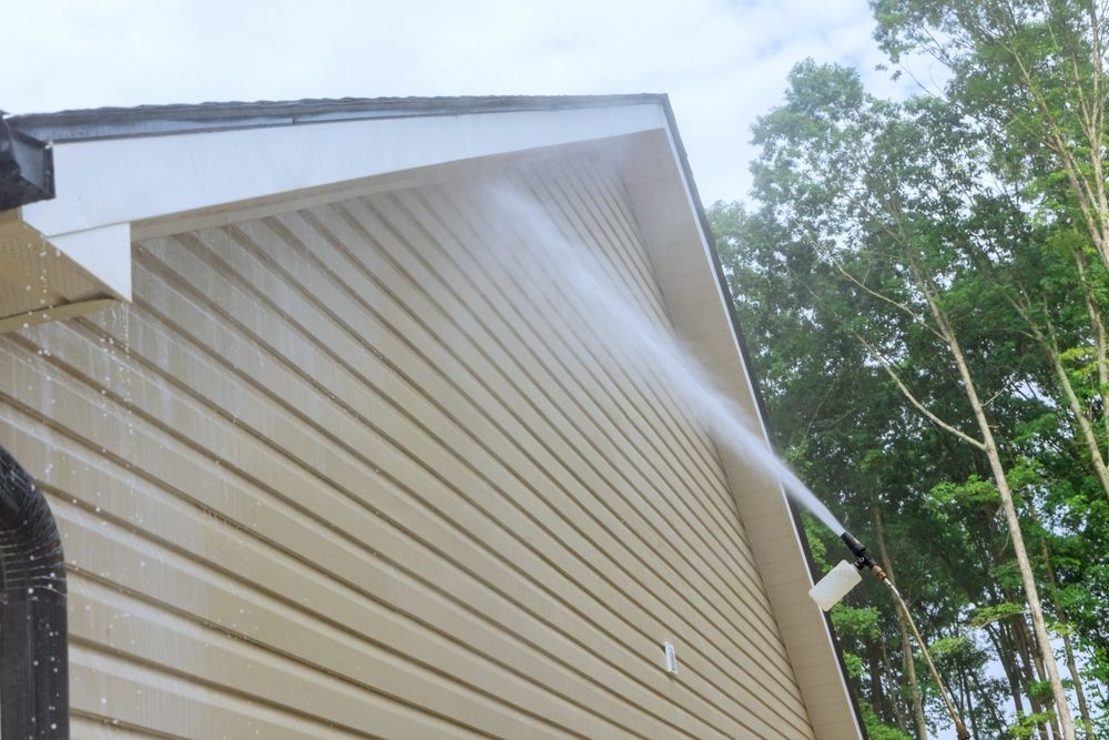 A pressure washer sprays a stream of water onto the beige vinyl siding of a house gable.