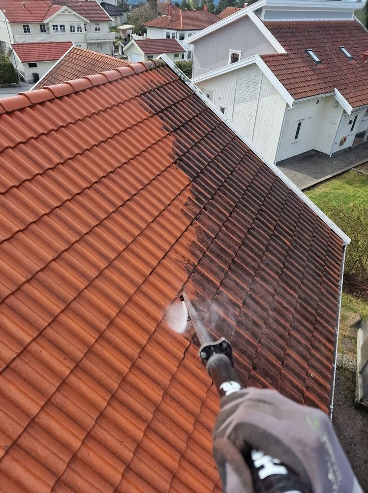 A person pressure washing a dirty, dark tiled roof, revealing a clean, bright orange surface underneath.