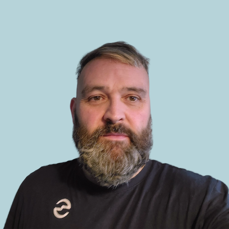 Man with a beard, in a dark shirt, against a blue background, looking directly at the viewer.