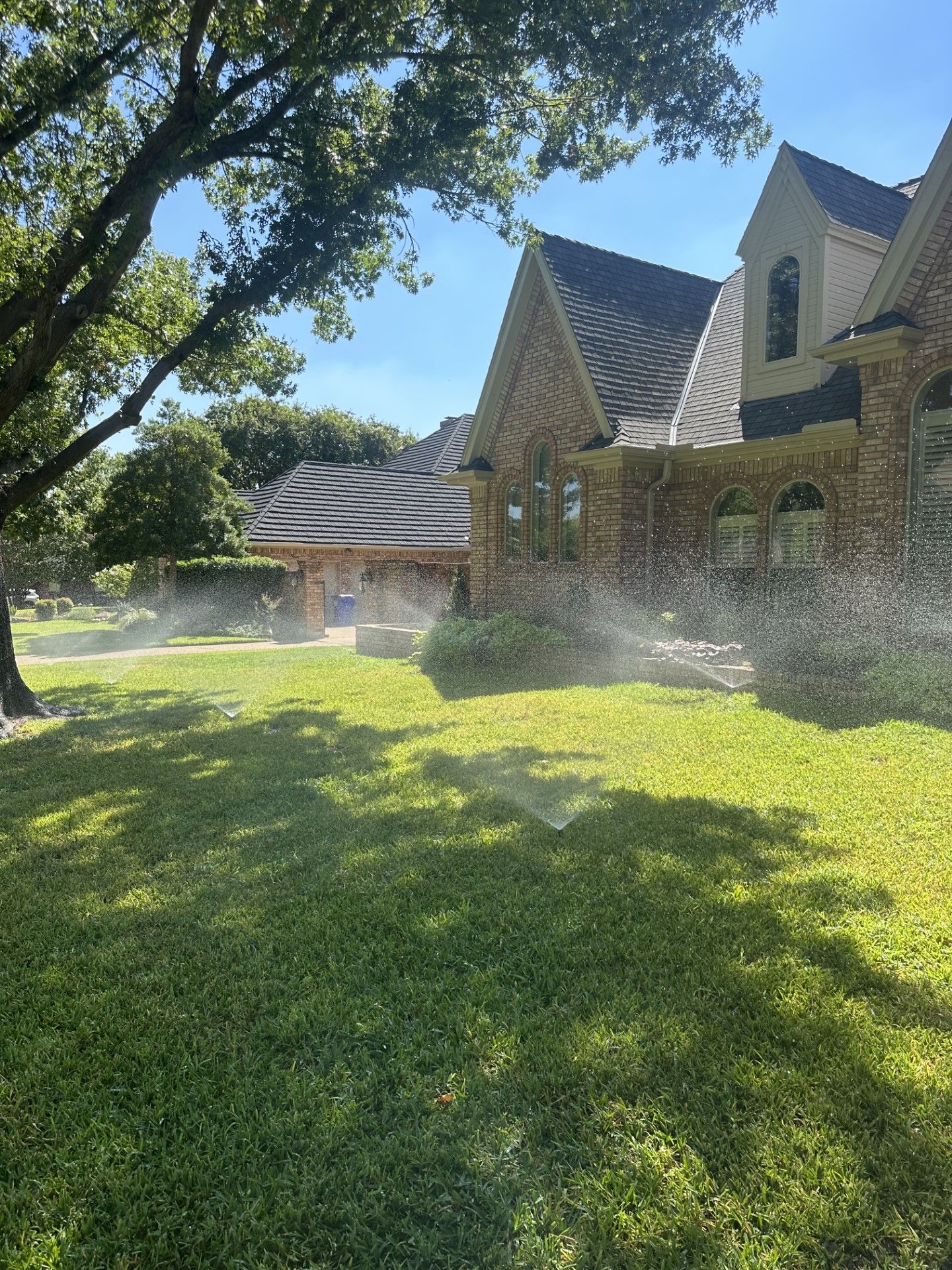 Lawn sprinkler watering green grass in front of a brick house with a blue sky background.
