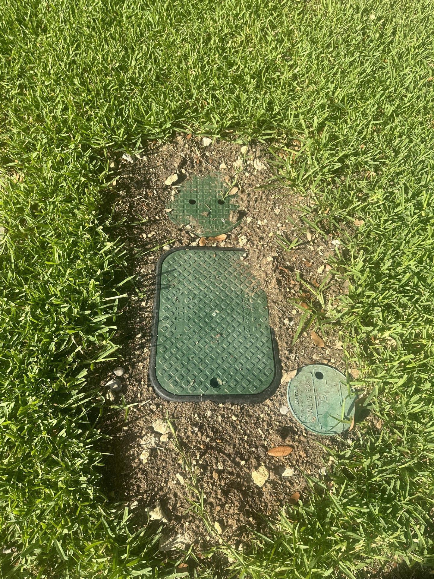 Green irrigation valve boxes in a patch of dirt surrounded by green grass.