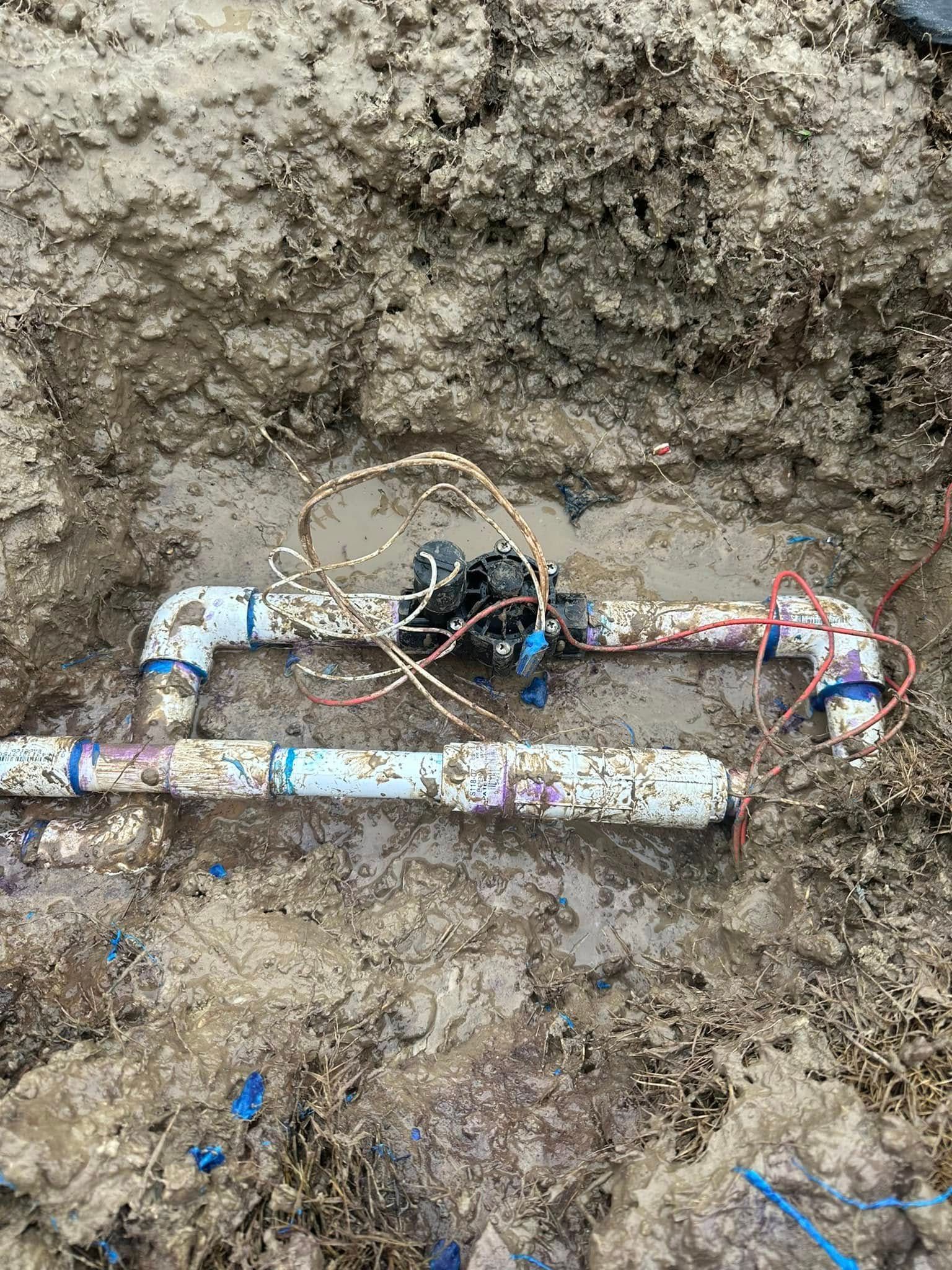 Pipes and valve in a dirt trench, possibly for irrigation or water supply, with exposed wires.