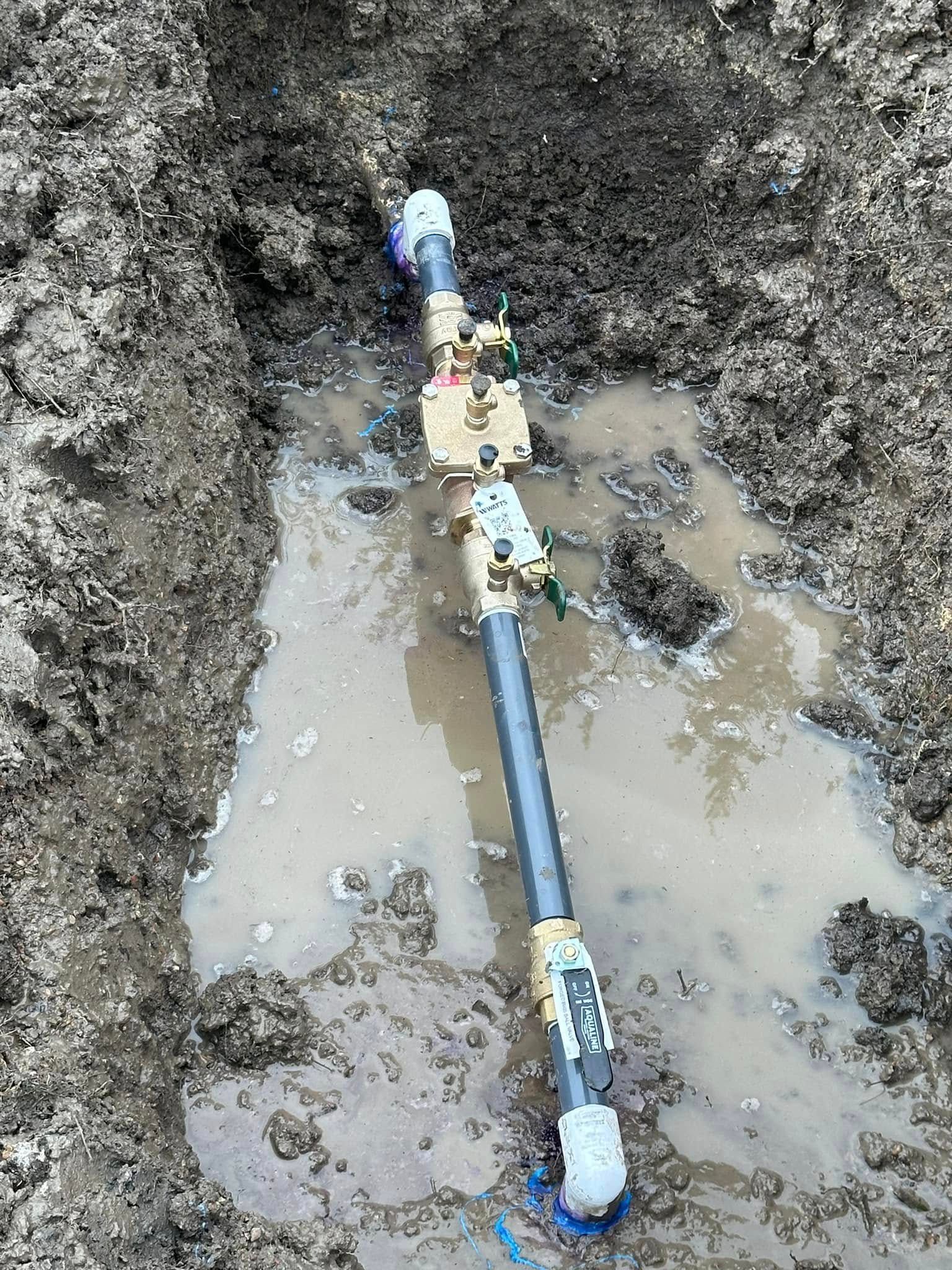 Plumbing repair in a muddy trench, showing pipes, valves, and connections with water pooling.