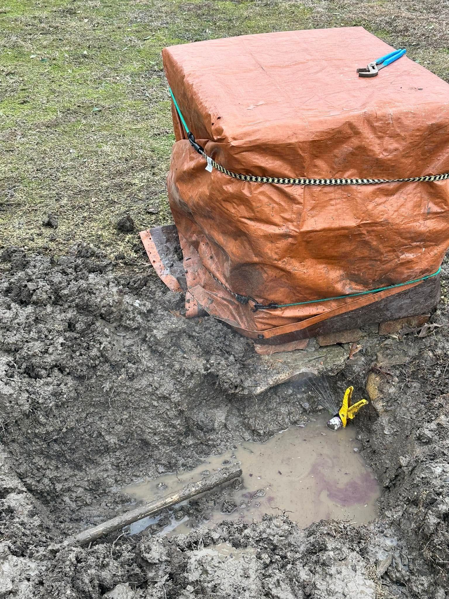 A muddy hole with water next to a covered brick structure and a utility knife in the muddy surface.