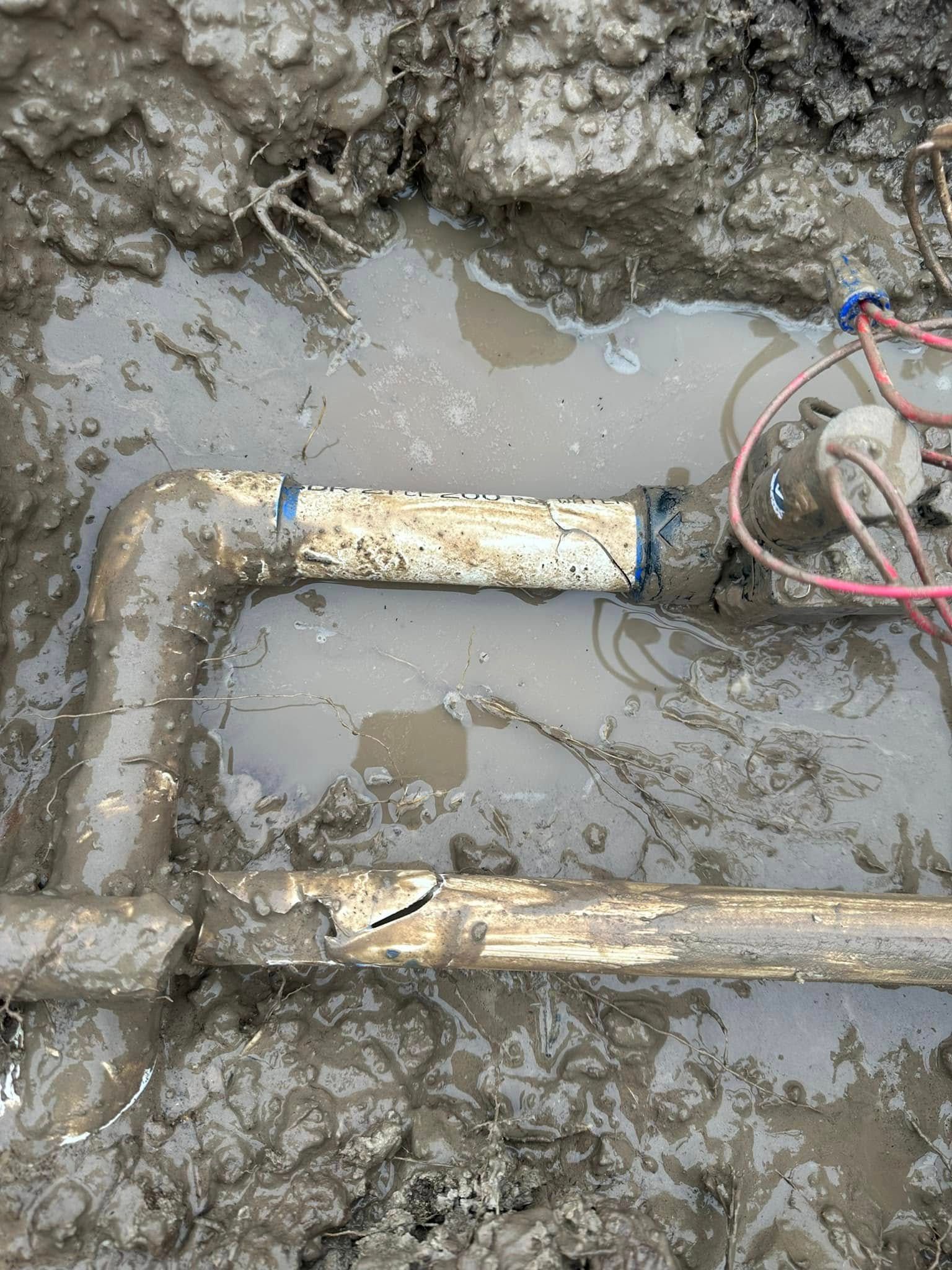 Muddy trench with broken PVC pipe, leaking water.