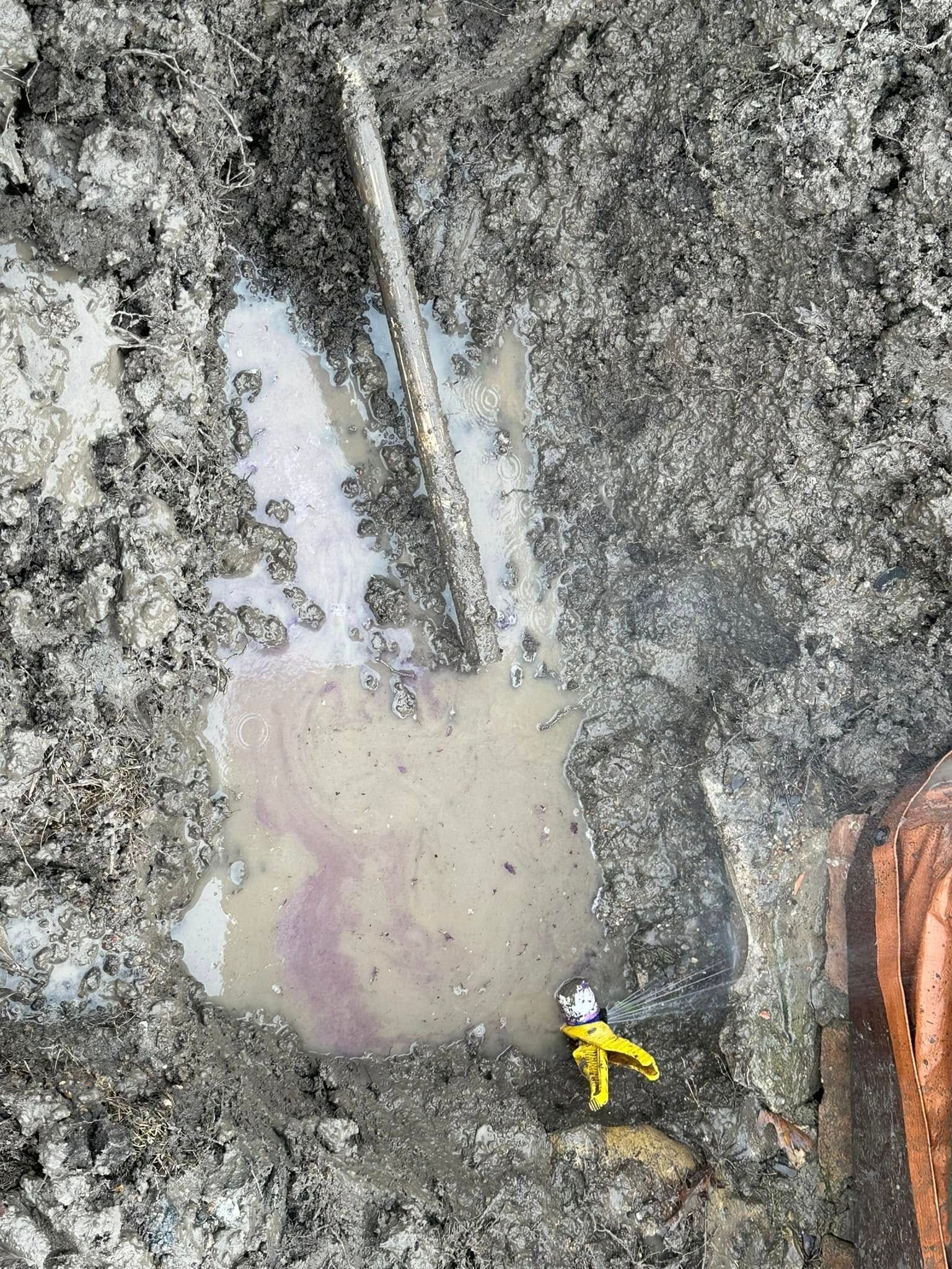 Pipe in muddy ground with a puddle of liquid, a worker in yellow protective gear at the bottom.