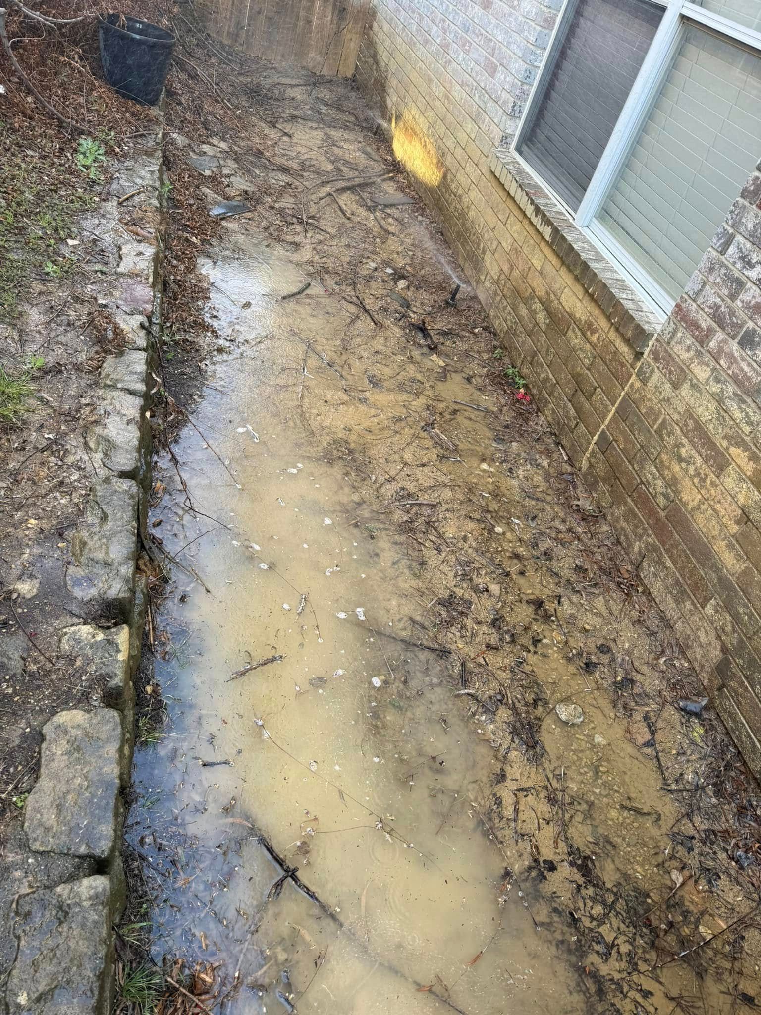 Muddy, debris-filled trench next to a brick building. Water pooled, near a window with blinds. Stone border on left.