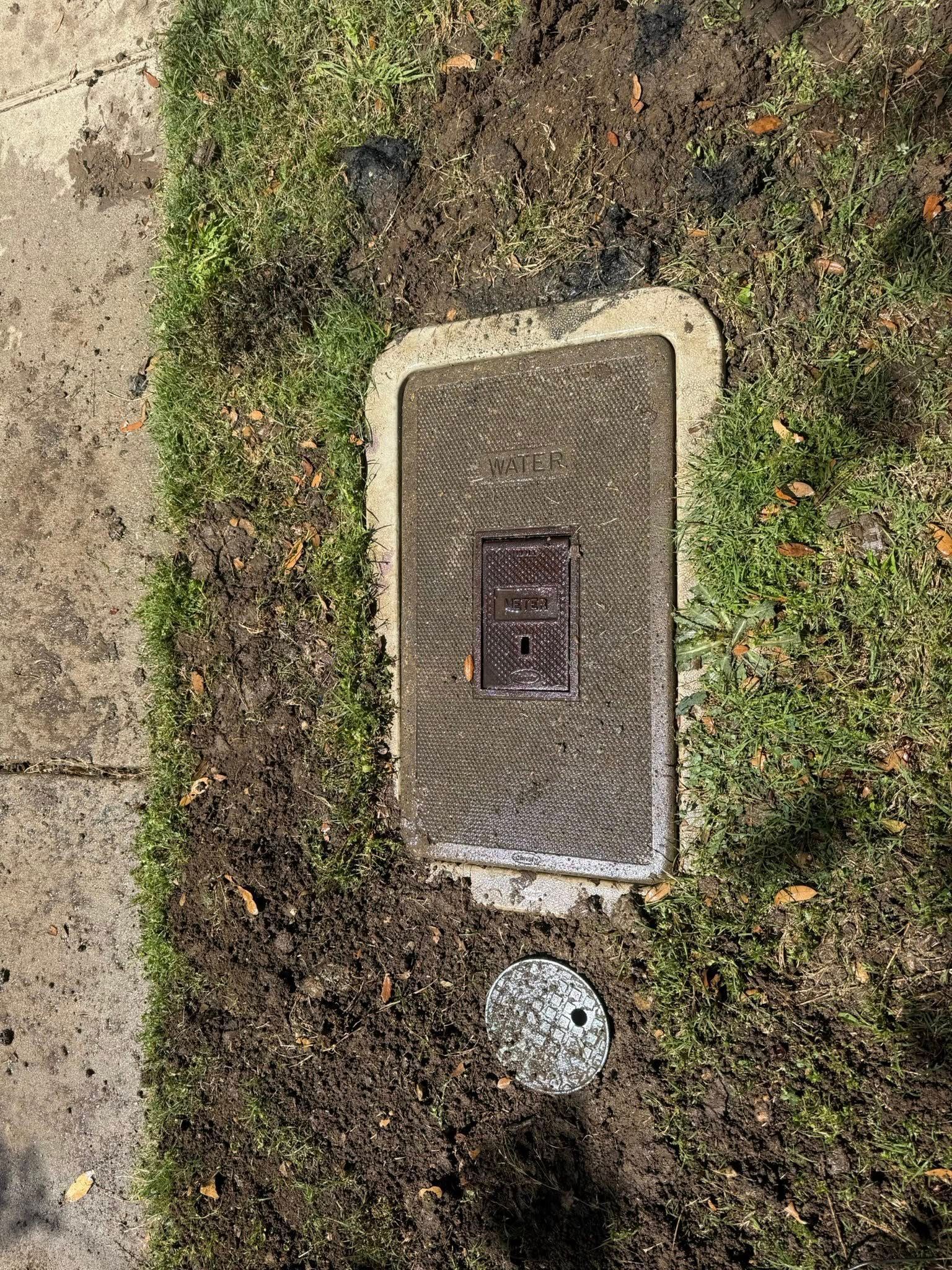 Rectangular utility box and circular cap in muddy grass. Concrete sidewalk to the left.