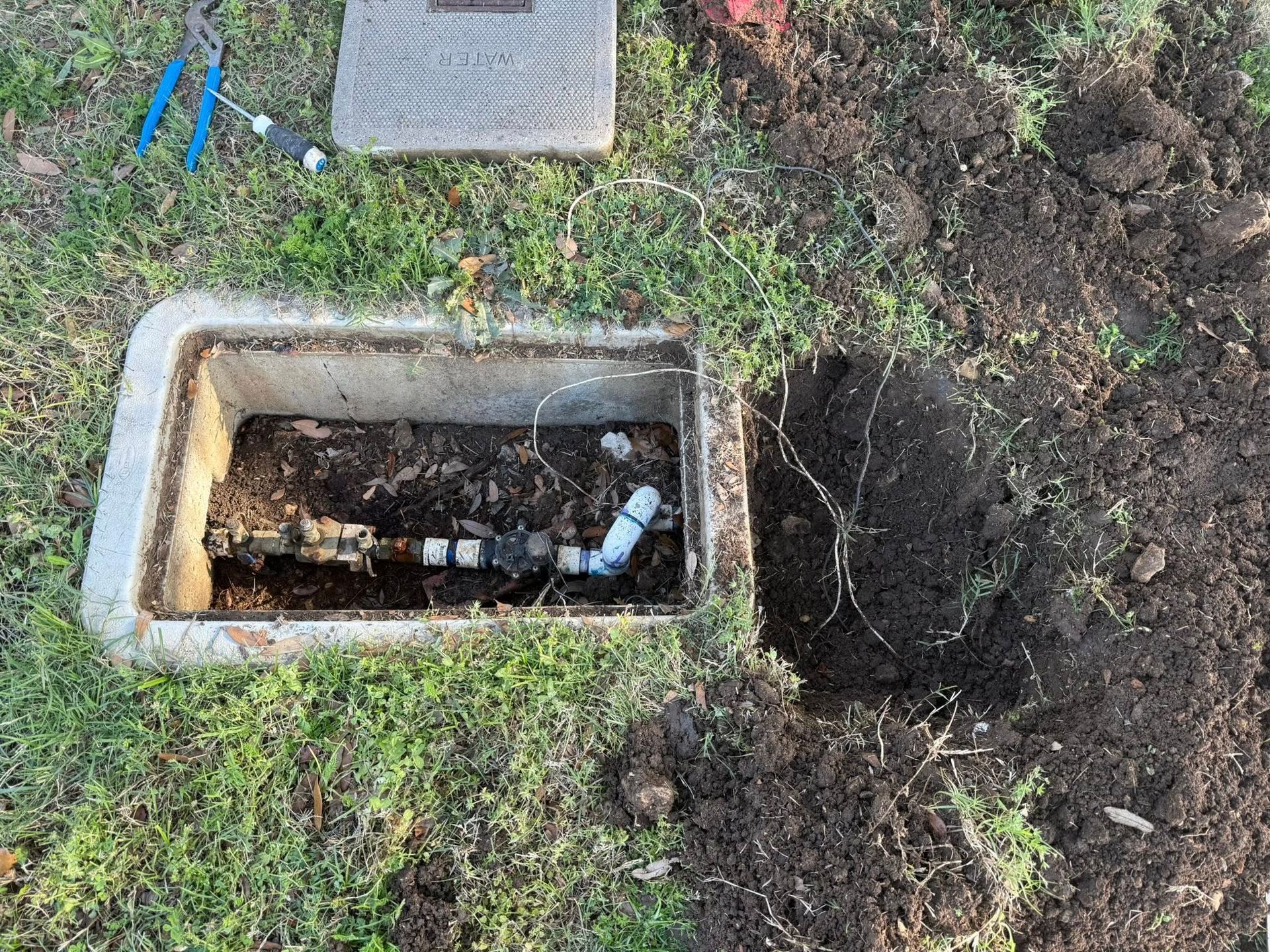 Open concrete box in grass with exposed sprinkler system pipes and dirt.