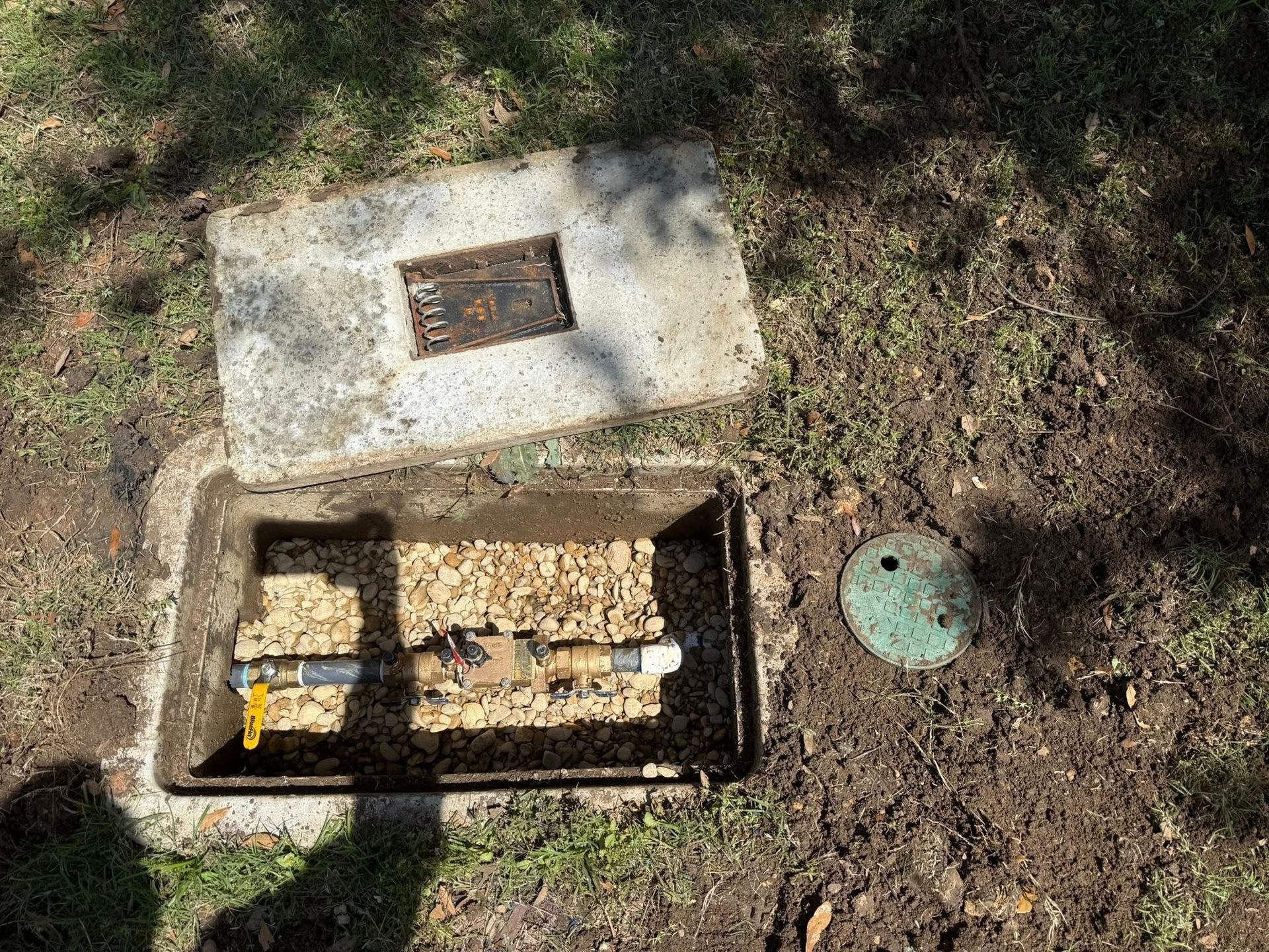 Open water meter box with a concrete lid on the ground. Inside are pipes and valves, covered with wood chips.
