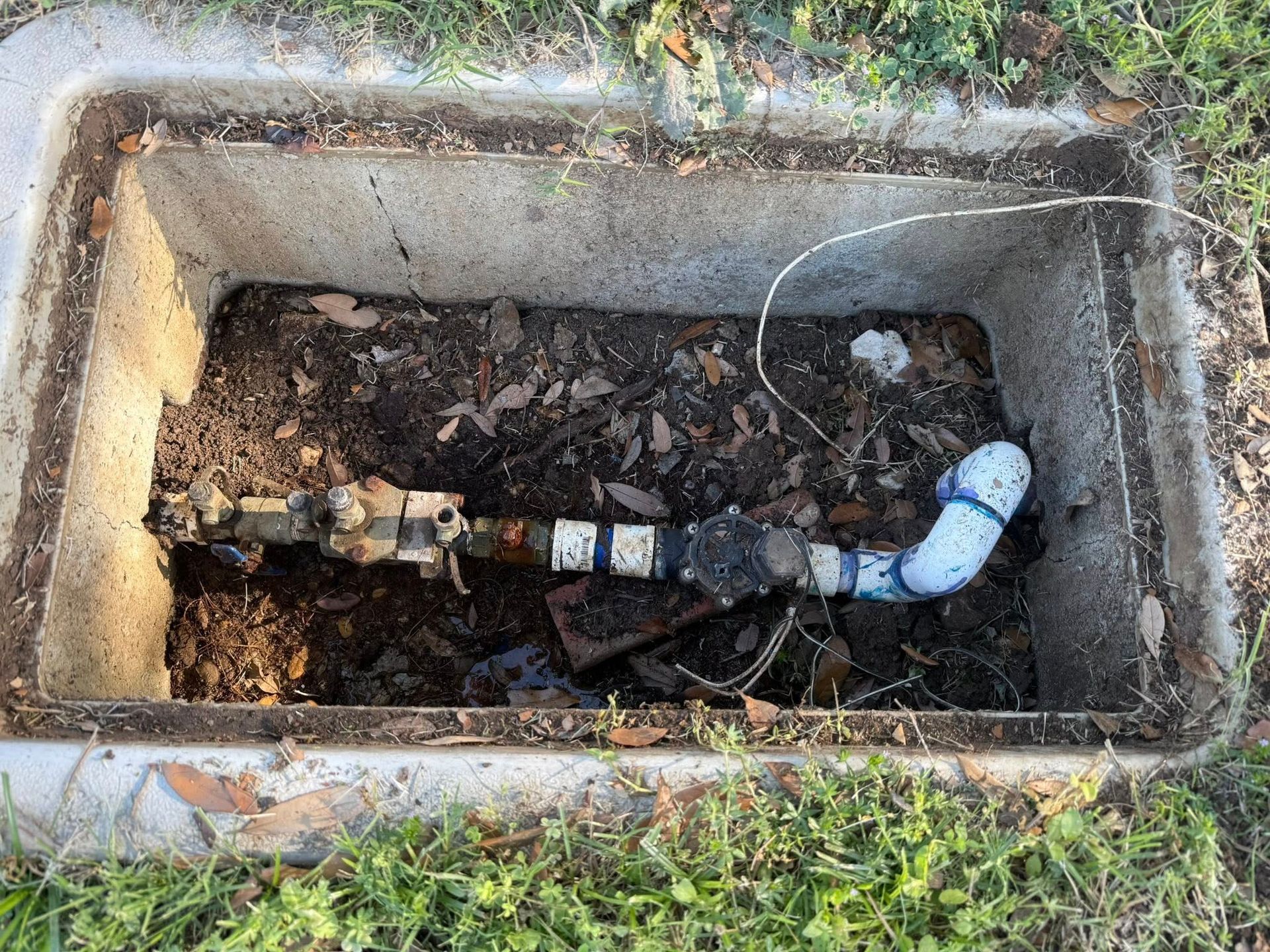 Water valve assembly inside a concrete box, surrounded by dirt and grass.