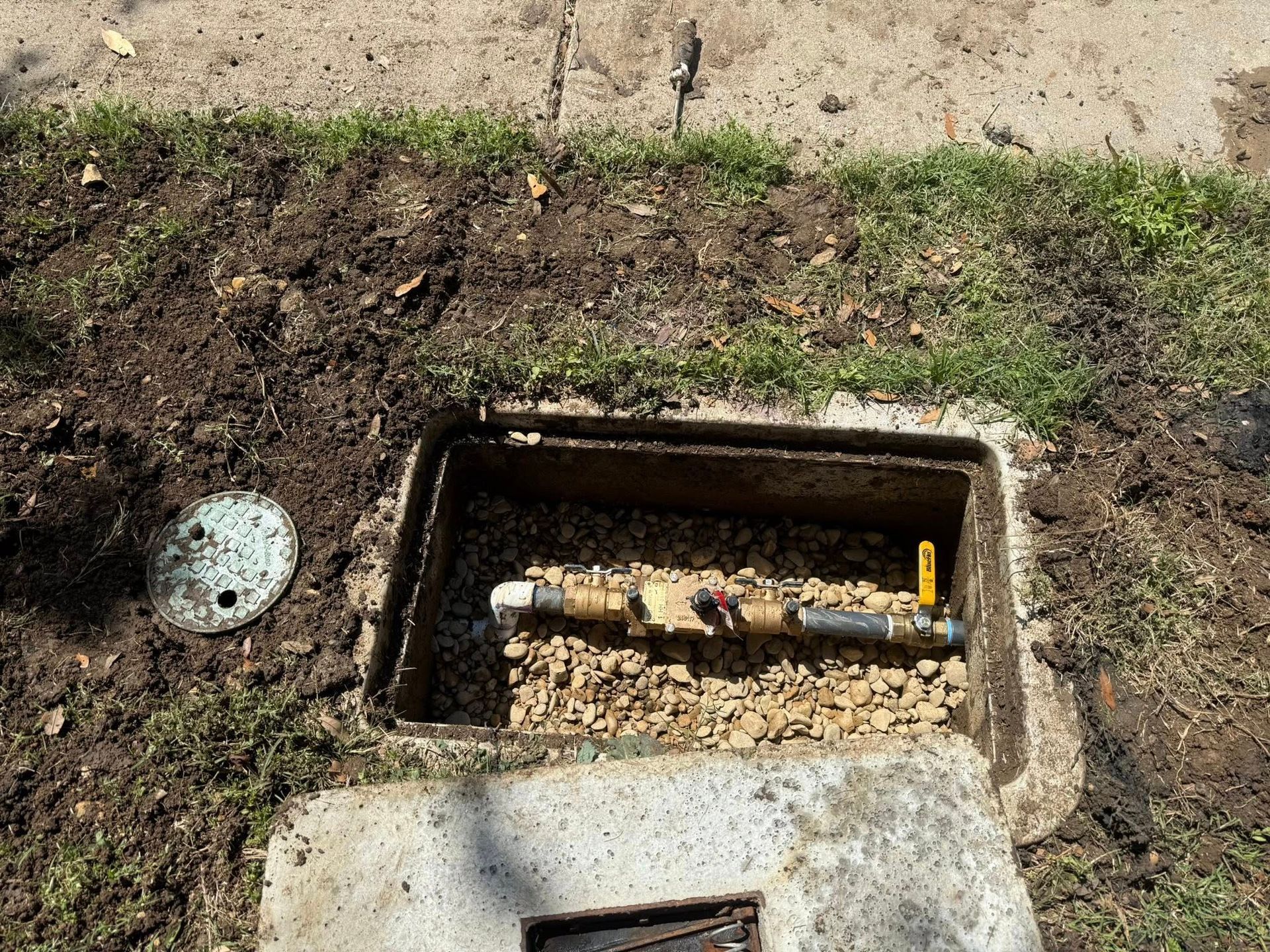 Open utility box with water meter and gravel base, near grass and sidewalk.