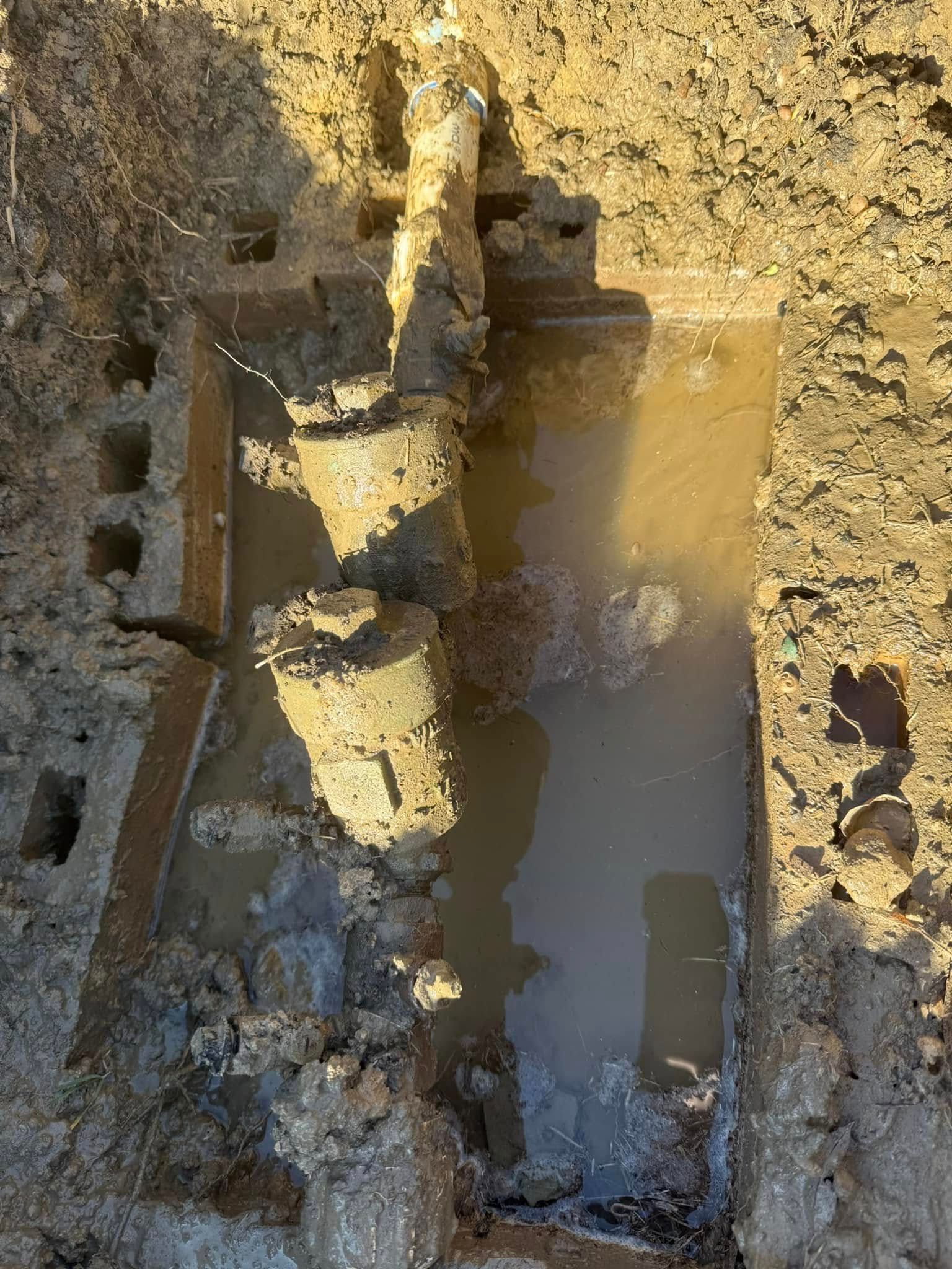 Excavation revealing buried pipes and structures in a muddy pit, filled with water under sunlight.