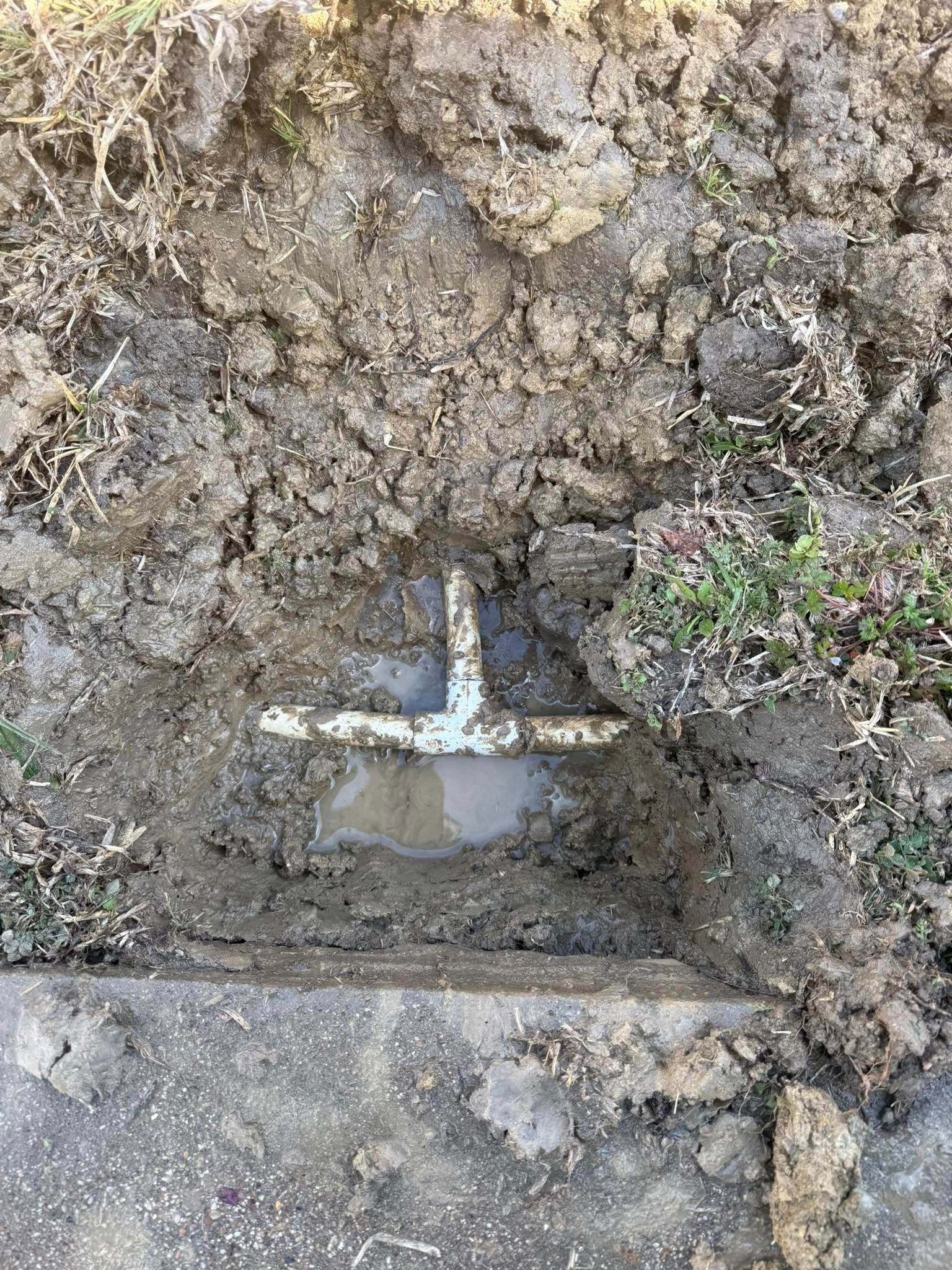 A buried, corroded pipe fitting in muddy ground; adjacent to a concrete slab.