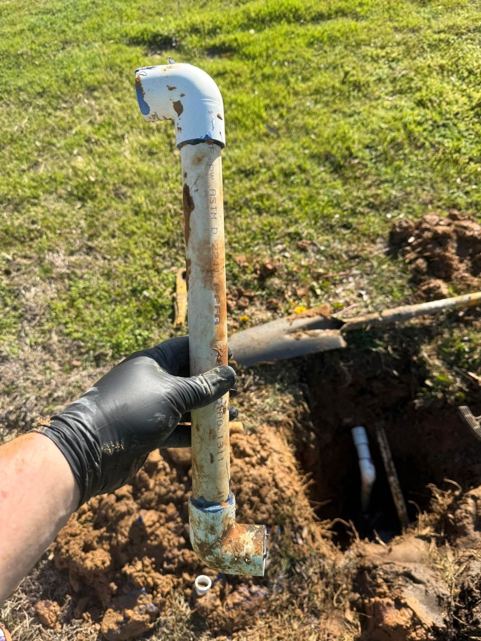 Hand holding a rusty pipe above a hole in the ground; white elbow joint at the top, grass in the background.