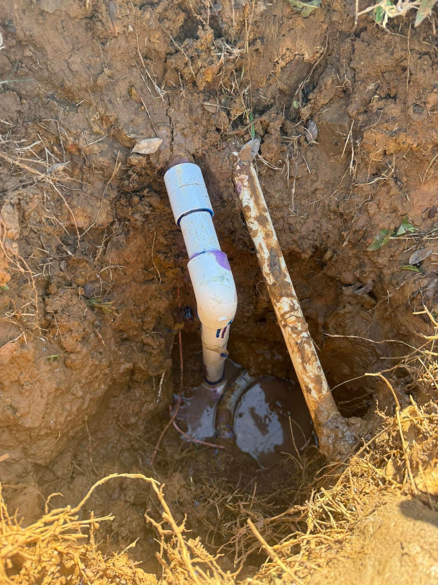 PVC pipe and fittings in an earthen hole, beside a metal pipe.
