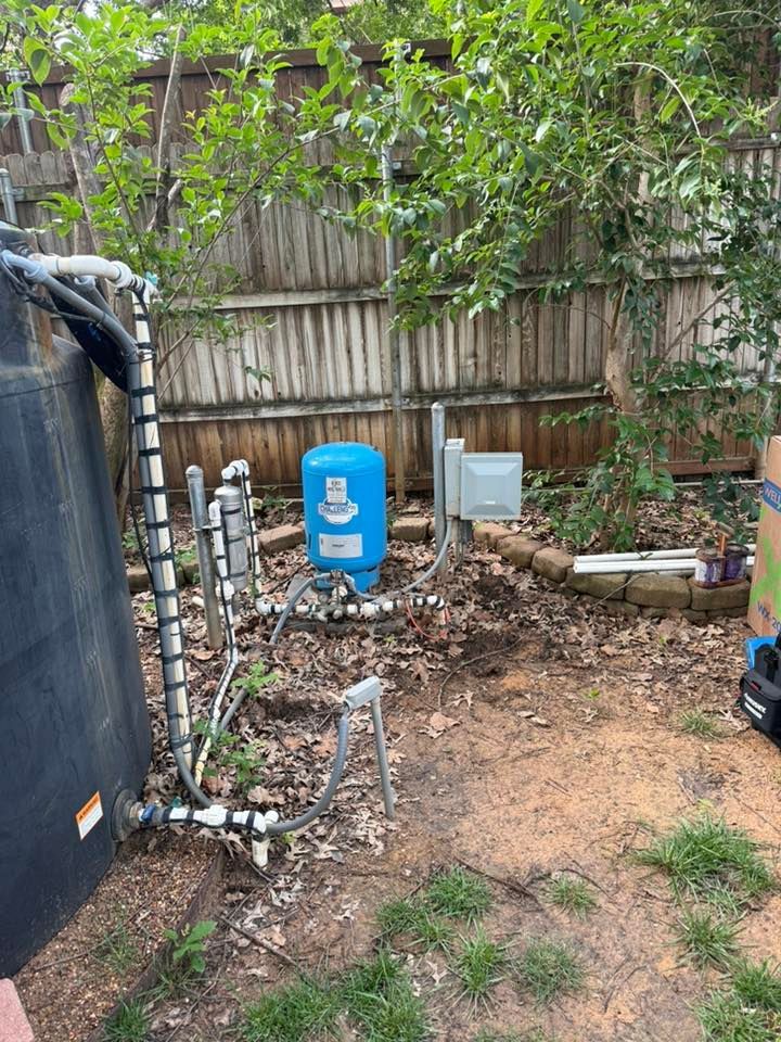 Backyard scene with a blue water tank, pipes, and electrical box against a wooden fence.
