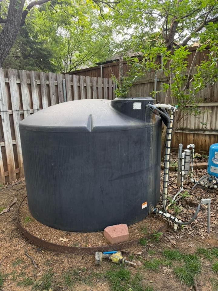 Large black water tank in a yard, near a fence and trees. Piping and valves are visible.