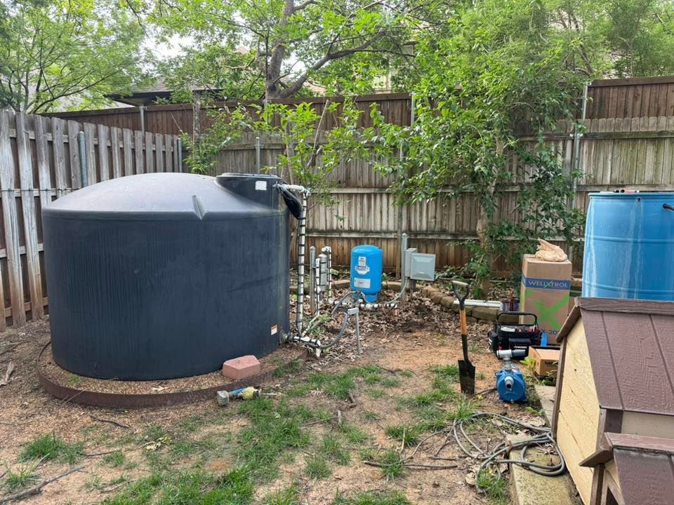 Black water tank in backyard with pipes, pump, and surrounding equipment.