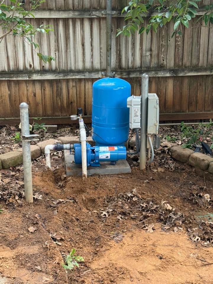 Well pump system outdoors with blue tank, motor, and electrical box near a wooden fence.