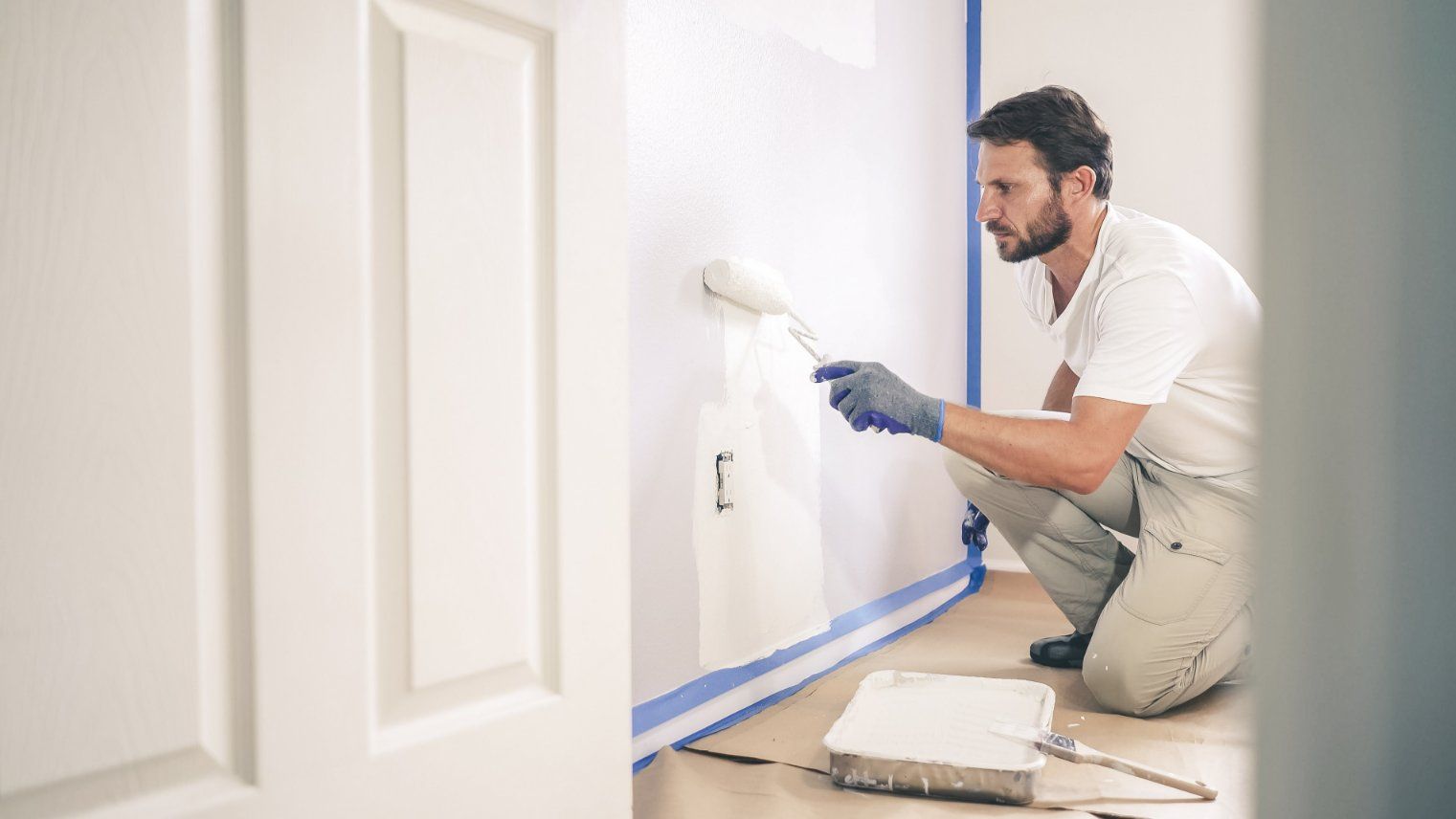Person painting a white wall with a paint roller, kneeling, wearing gloves.