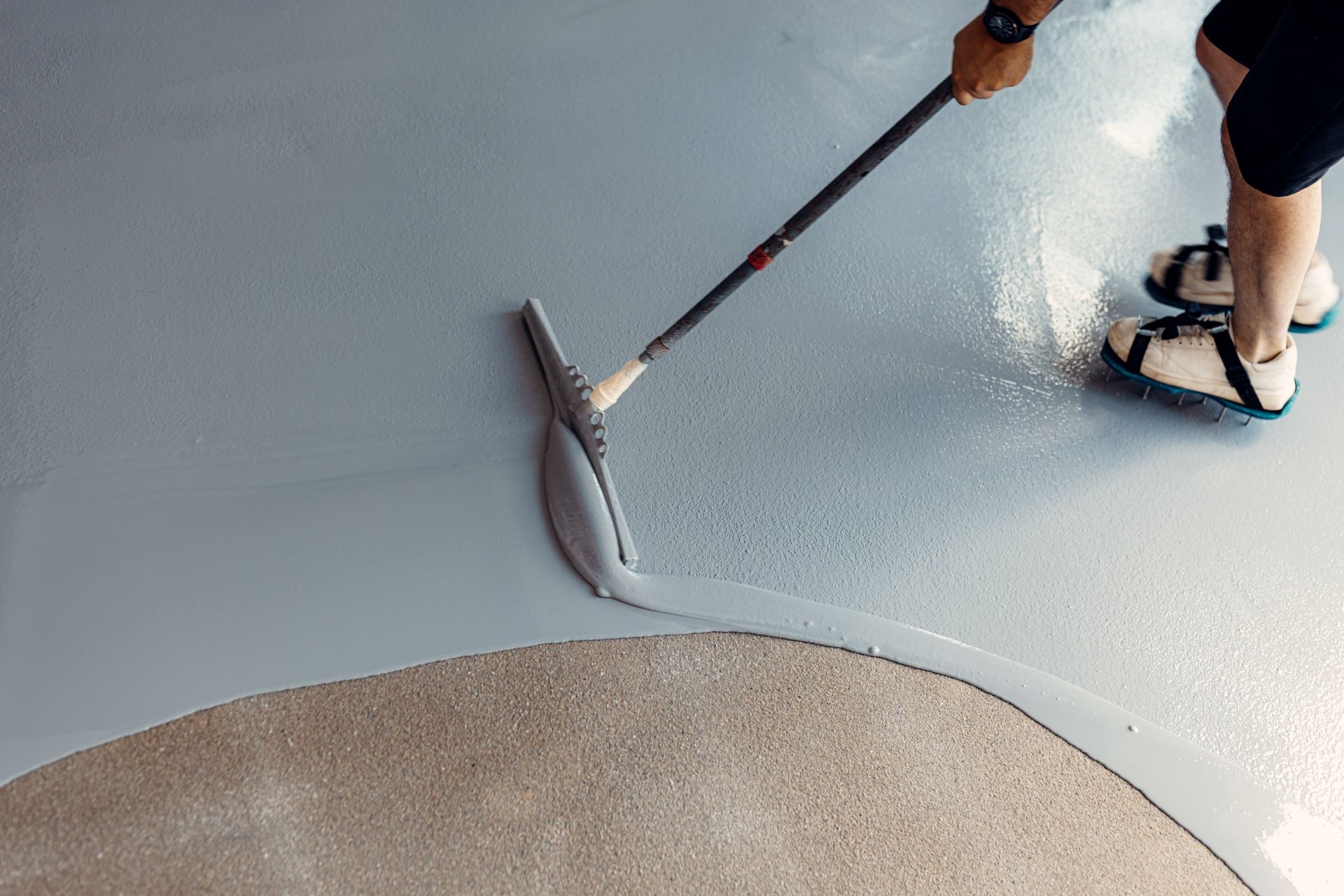 Person applying light gray epoxy coating on a garage floor with a squeegee.
