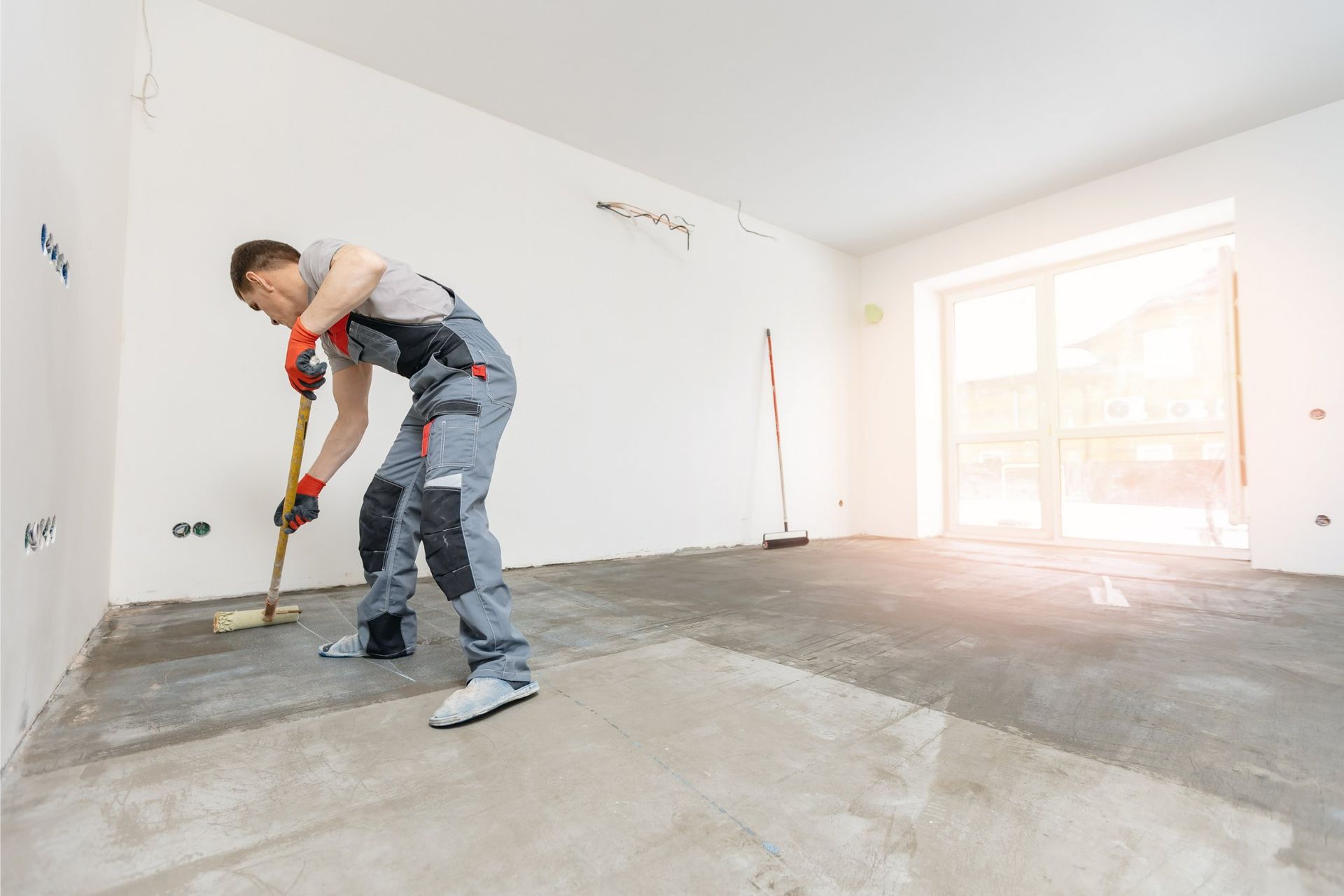 Worker smoothing concrete floor with a tool in an empty room, wearing work overalls and gloves.