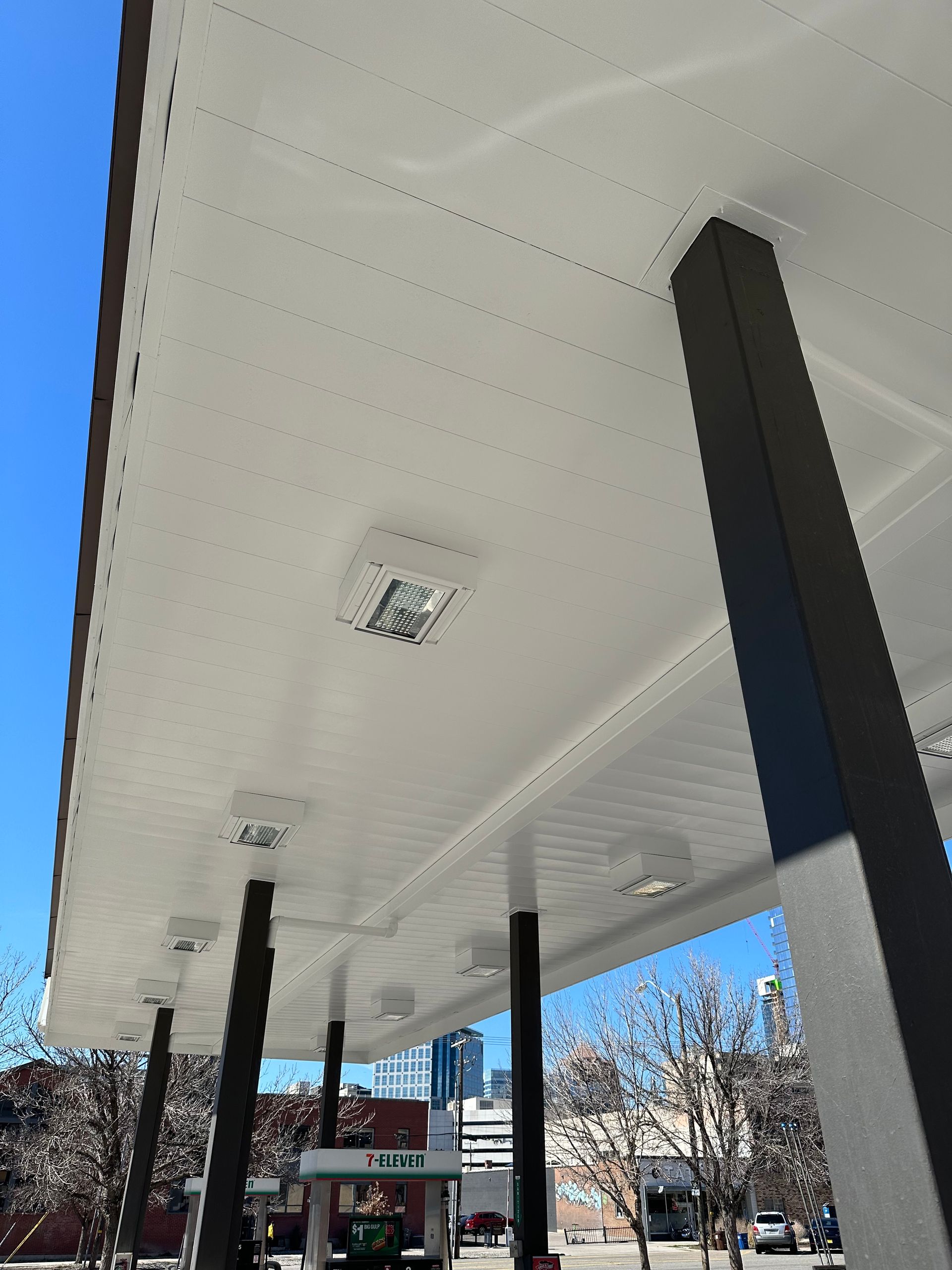 View from under gas station canopy, white ceiling, black support beams, clear blue sky.