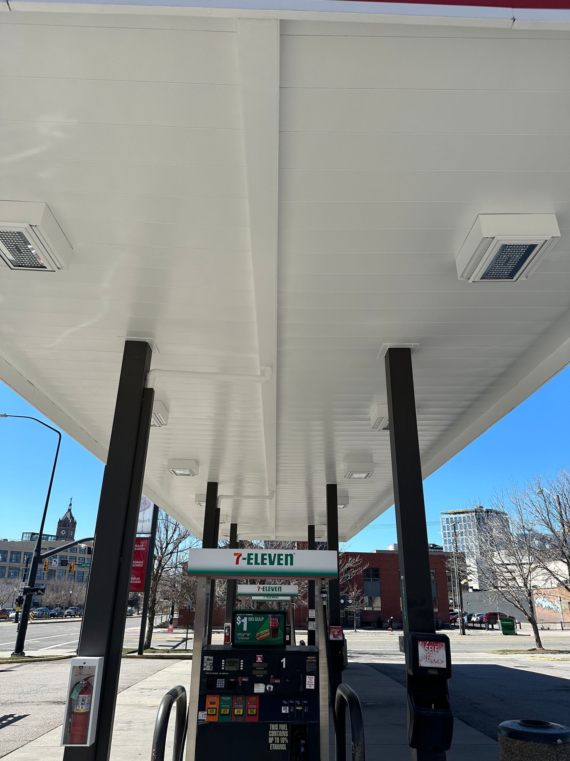 View from under a gas station canopy. A 7-Eleven gas pump is visible, set against a city skyline on a sunny day.