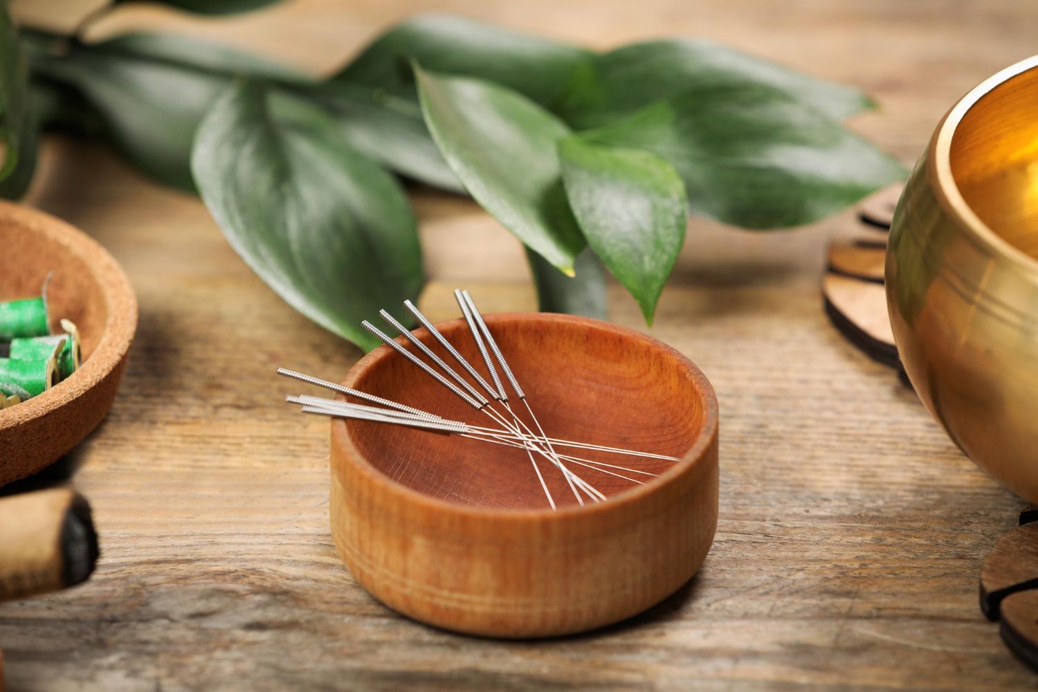 Copper bowl with acupuncture needles on a wooden table beside green leaves and a brass cup
