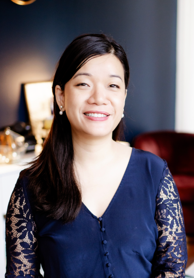 Smiling woman in a navy dress with lace sleeves standing indoors in front of a softly lit background