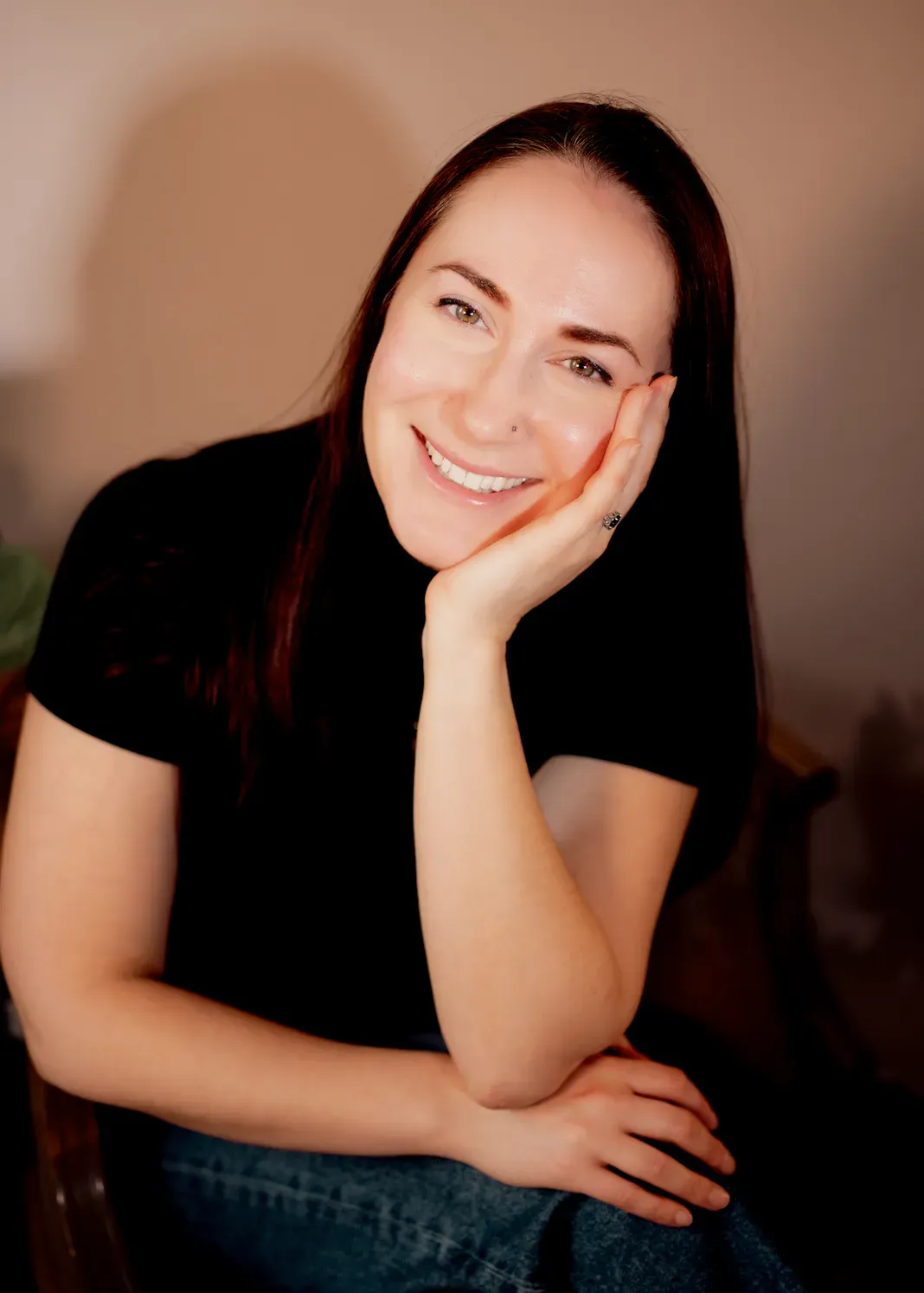 Smiling person with long dark hair rests their head on their hand against a warm indoor background.