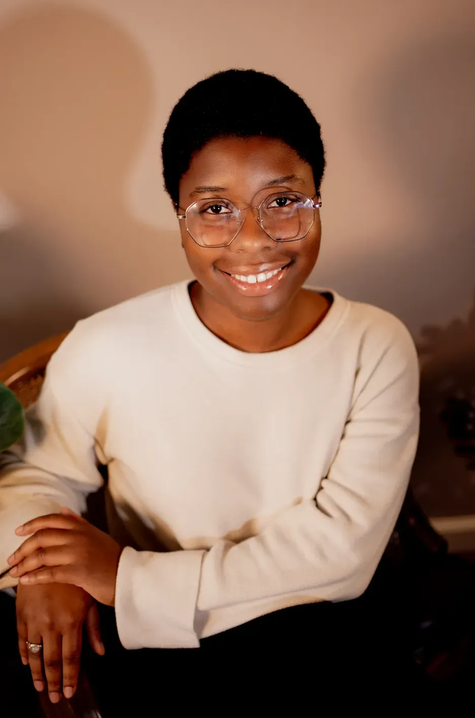 Smiling person in a cream sweater and glasses, seated against a warm brown background.