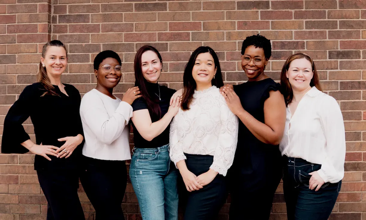 Six people posing together in front of a brick wall, smiling and standing side by side.