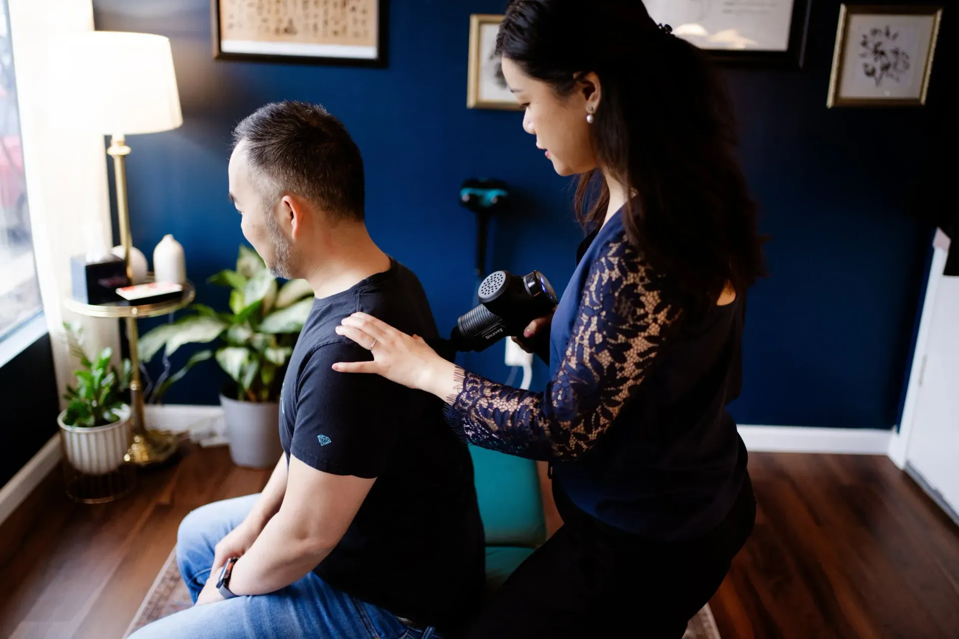Massage therapist working on a seated client in a dim room with blue walls and plants