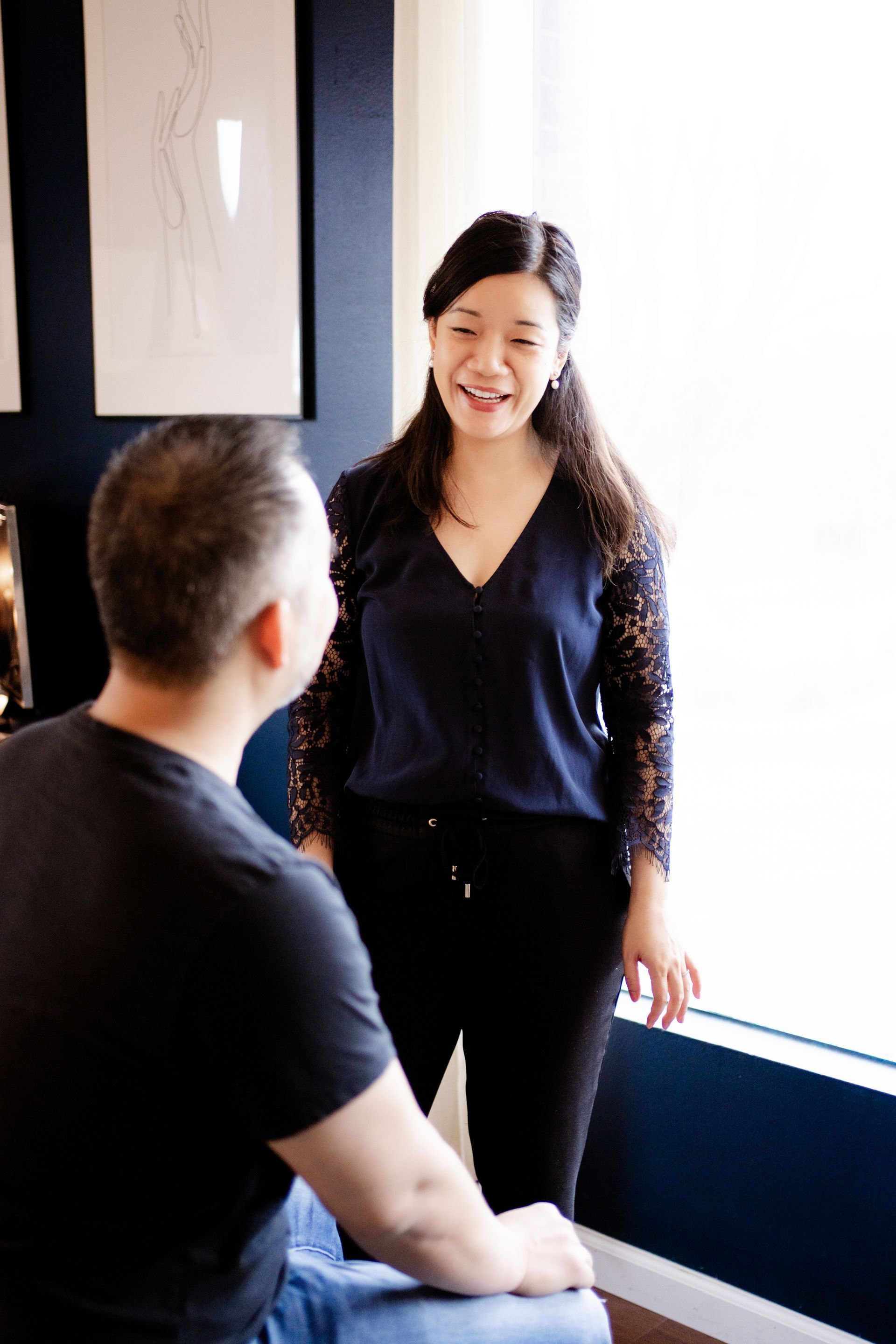 Two coworkers talking in a bright office, one standing and smiling while the other sits facing her.