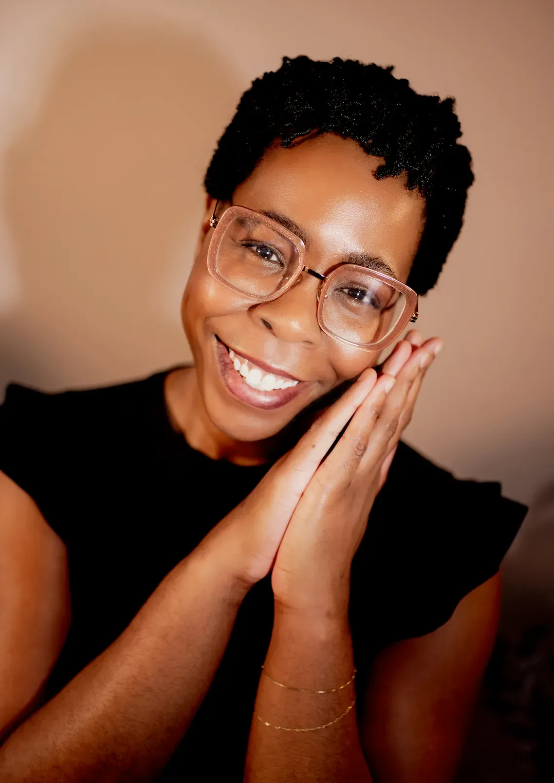 Smiling person with glasses resting their face on both hands against a warm tan background