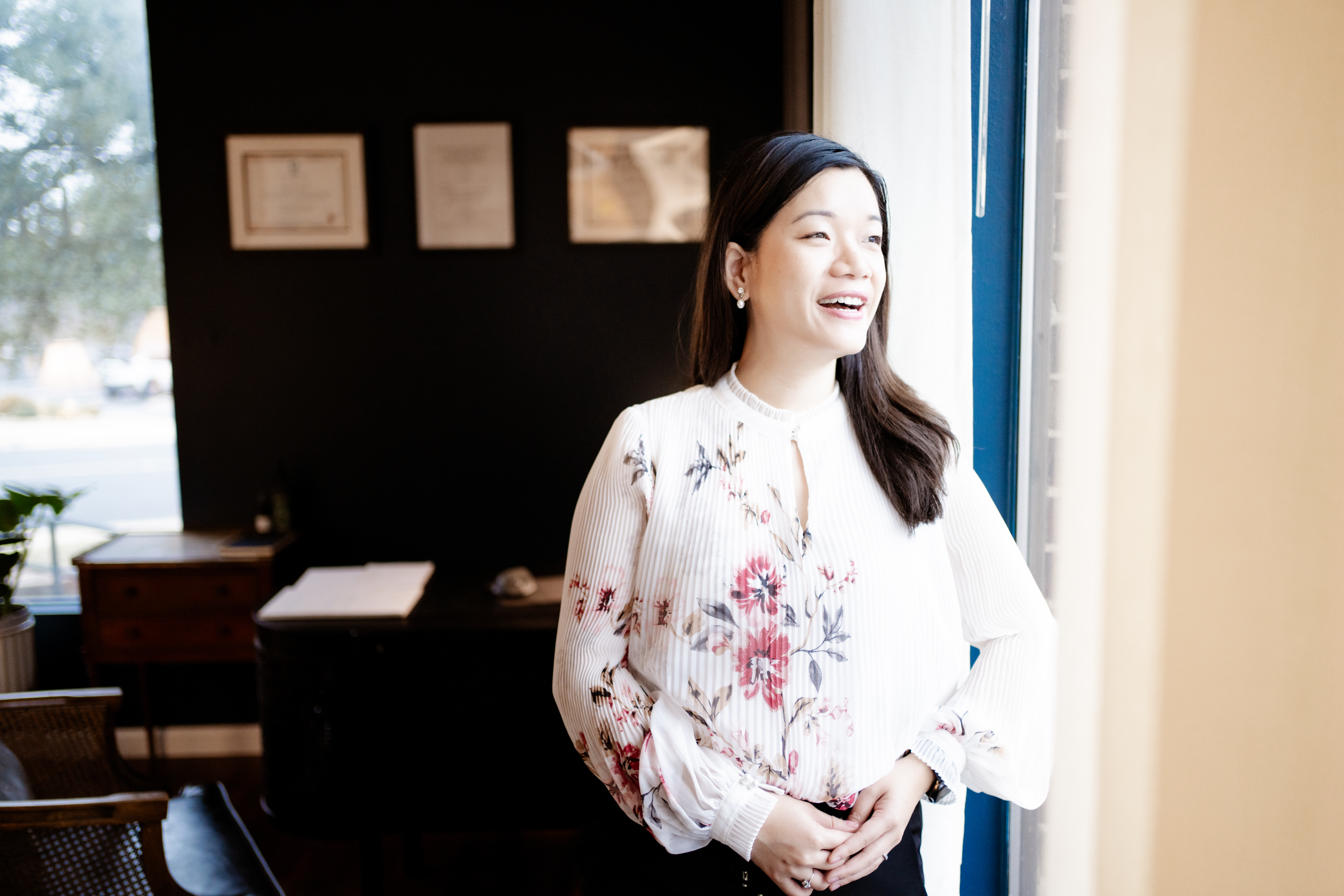 Woman in a floral blouse standing by a window, smiling in a softly lit room.