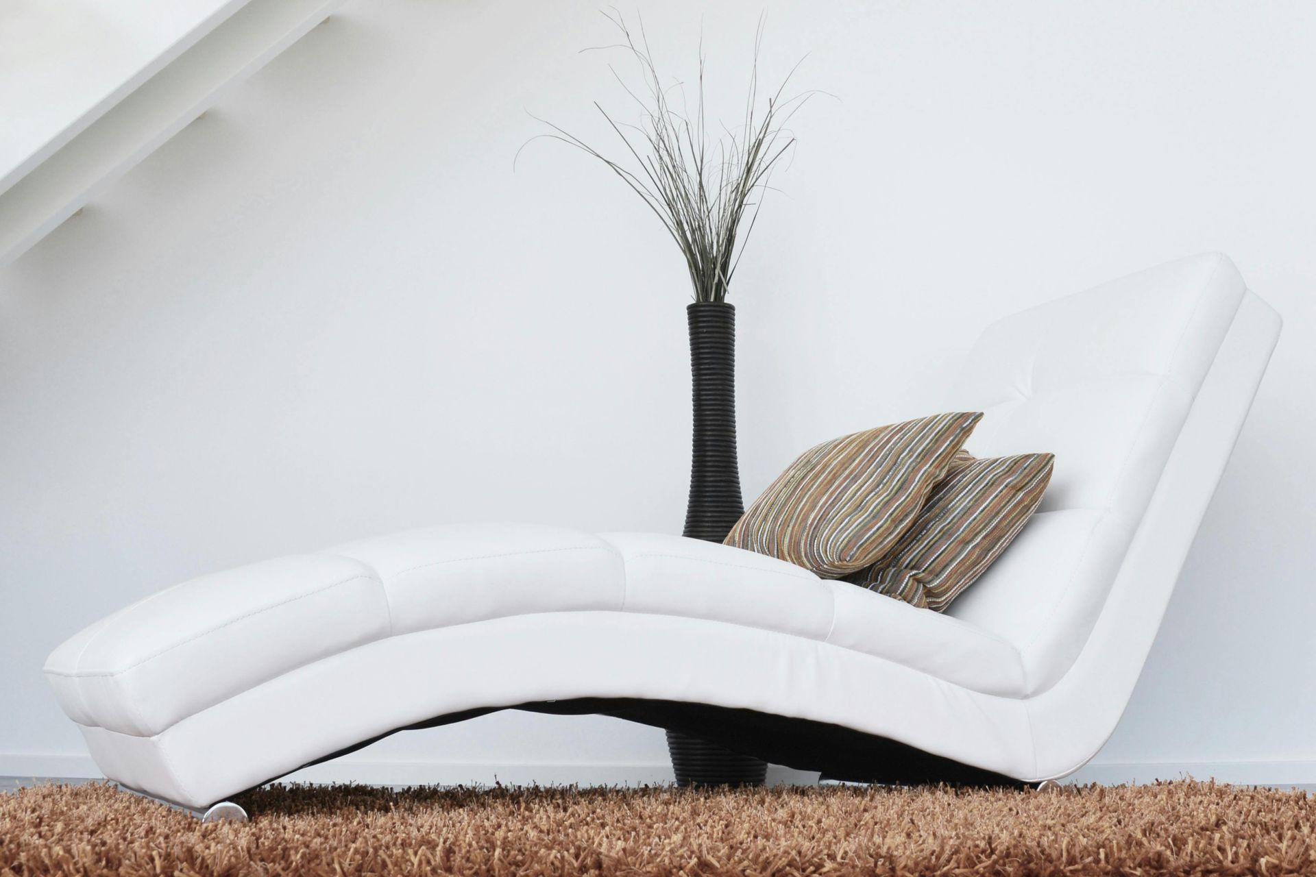 Chaise longue bianca moderna con cuscini a righe e un alto vaso nero su un tappeto a pelo lungo.