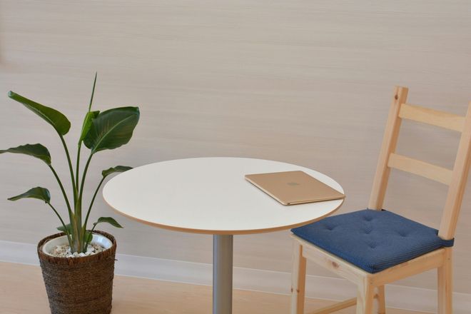Un tavolino rotondo bianco con un quaderno, accanto a una pianta in vaso e una sedia di legno in una stanza minimalista.