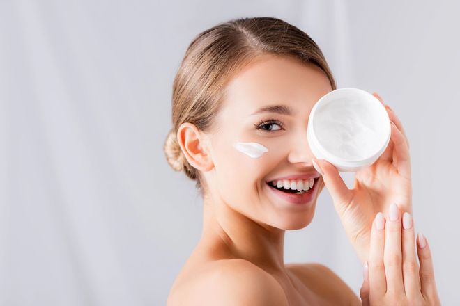 Una persona sorridente si applica una crema per il viso mentre tiene in mano un vasetto bianco, su uno sfondo grigio chiaro.