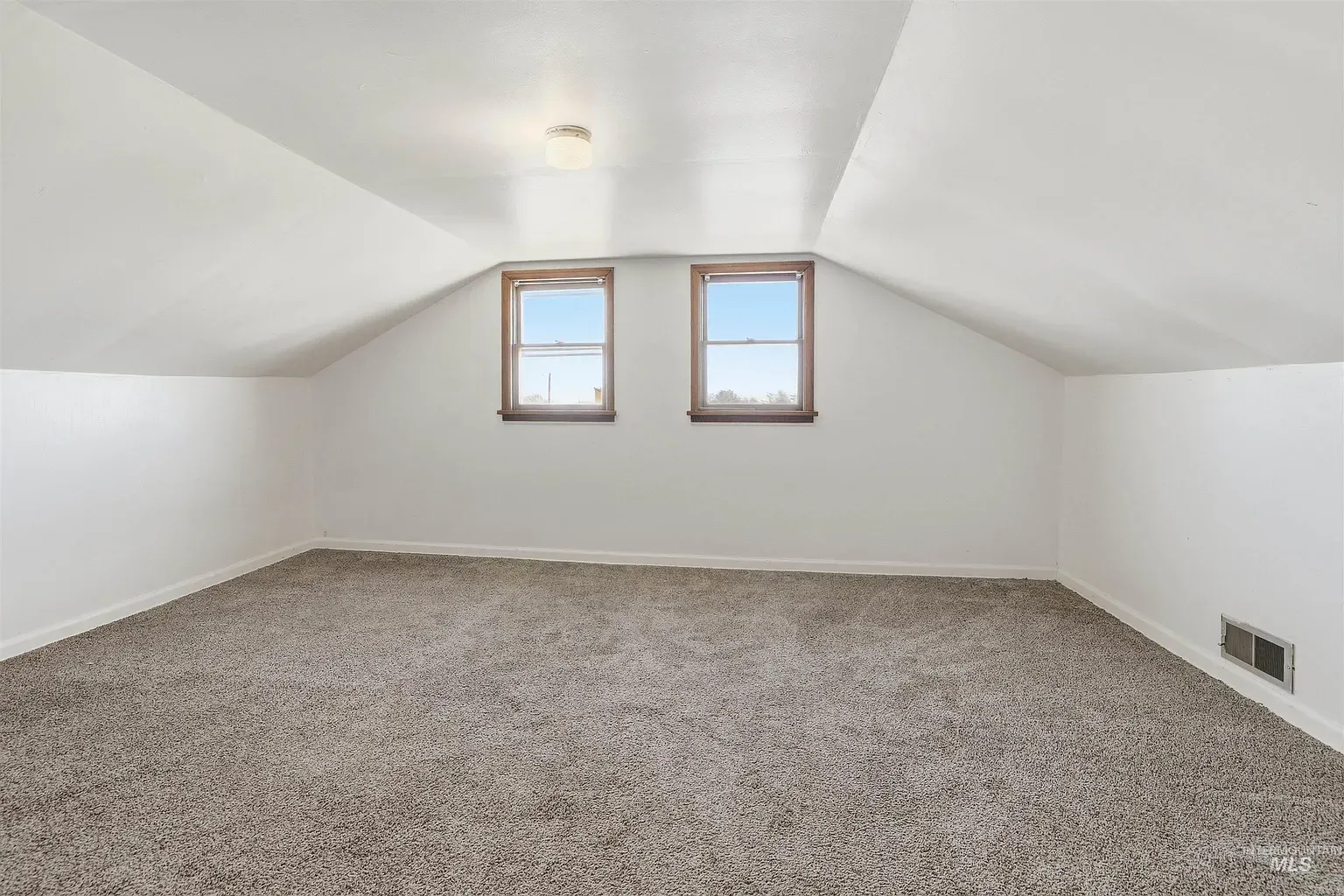Empty carpeted attic room with sloped white walls and two small windows.