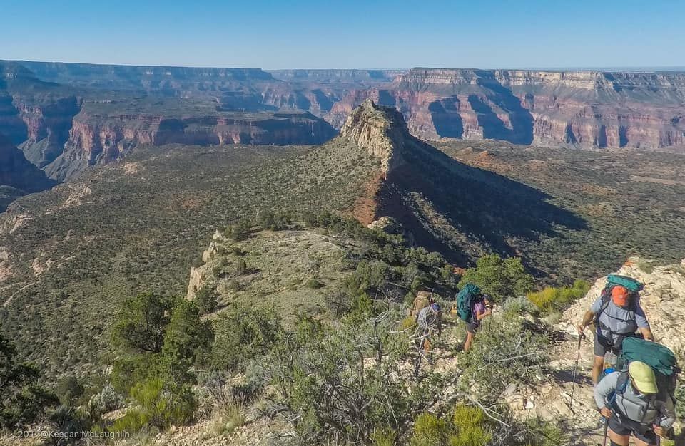 Hikers on a rocky canyon trail overlooking a vast red-and-green canyon landscape