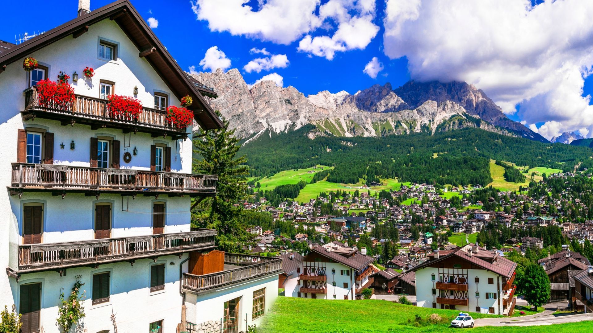 Alpine village with chalet balconies, green hills, and dramatic rocky mountains under a bright blue sky